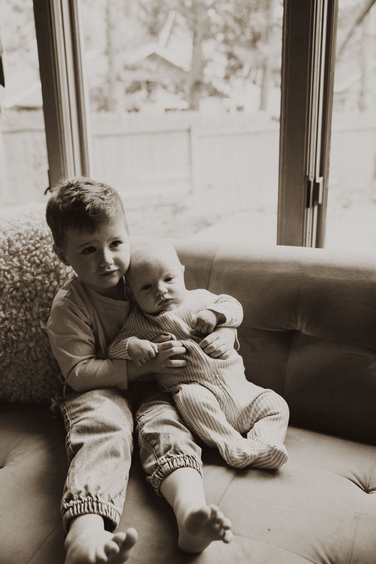 Black and white photo of older boy hugging baby sibling on green couch near window.