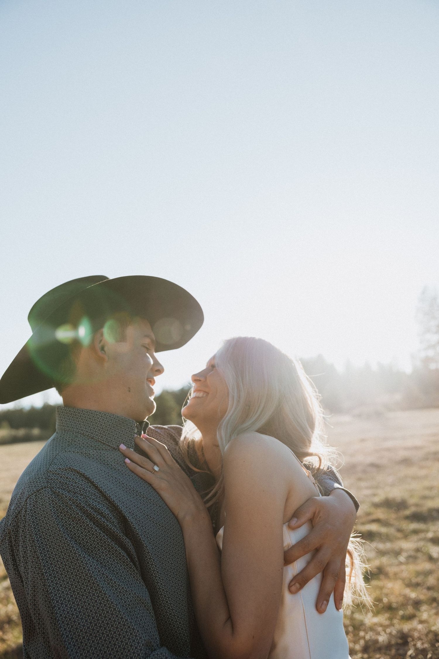 A couple embraces and laughs together in a sunlit field, man wearing cowboy hat and sunglasses.