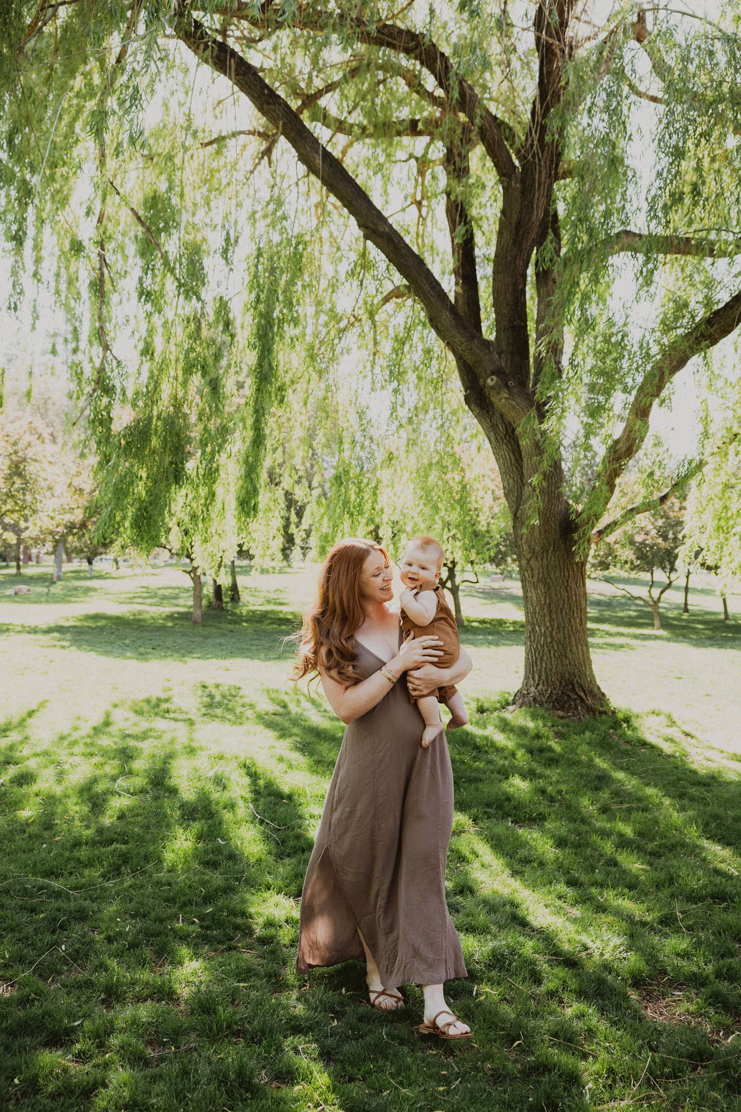 A mother in a mauve dress holds her baby under a weeping willow tree in a lush green park.