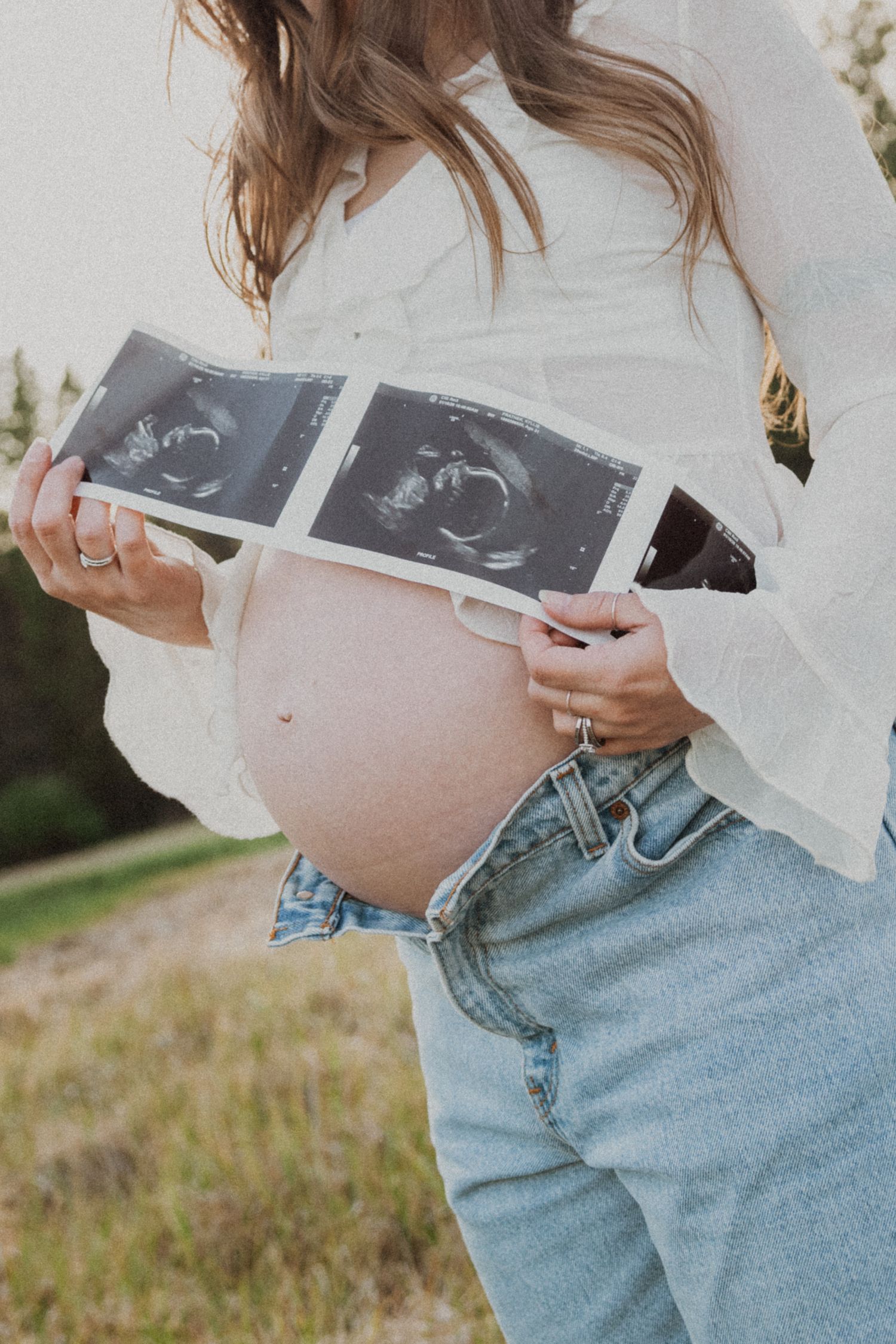Pregnant woman in white shirt and jeans holding ultrasound photos over baby bump outdoors.
