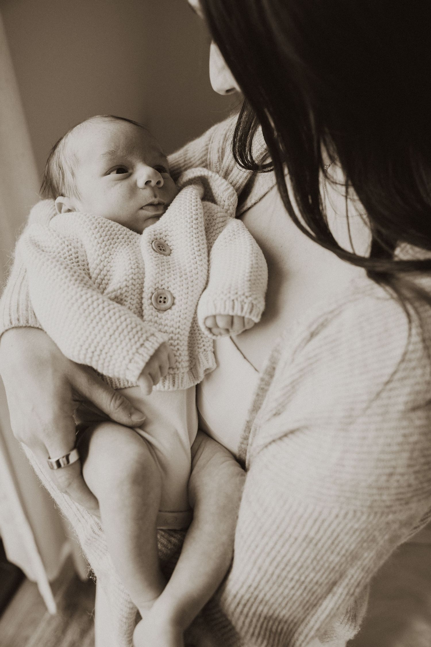 Close-up sepia photo of a newborn baby in a knit cardigan being held by their dark-haired mother.