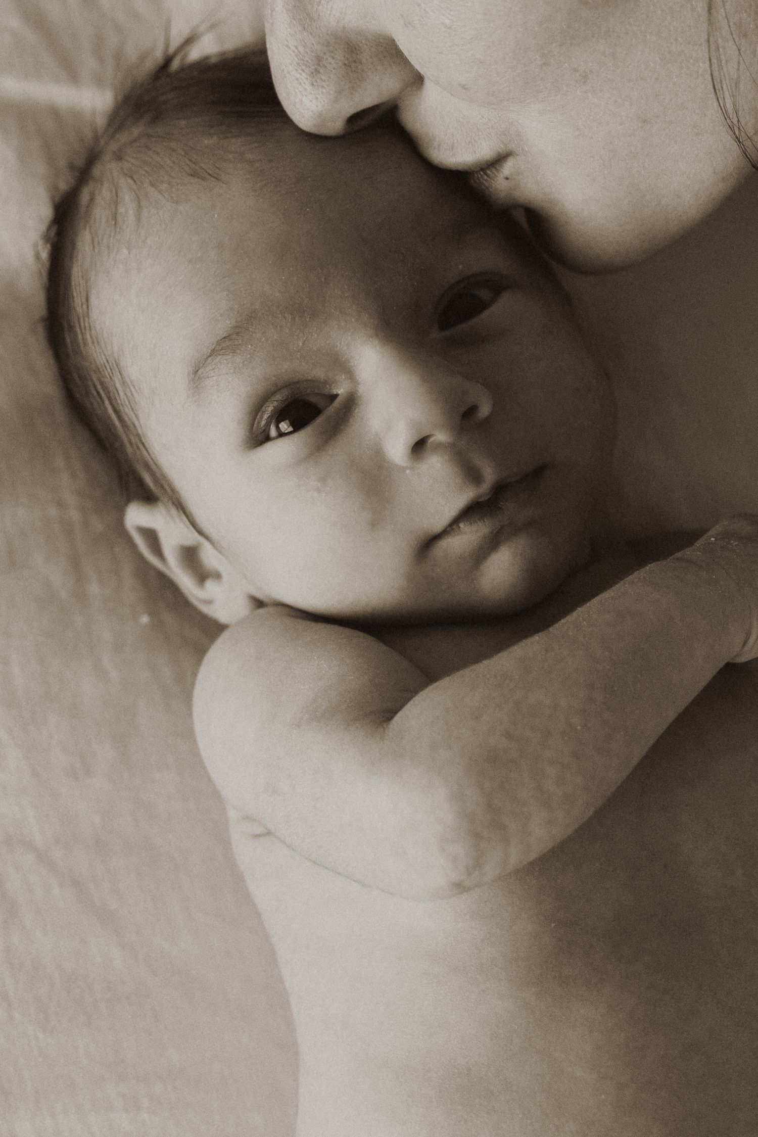 A mother tenderly kissing her newborn baby's head in a soft sepia-toned portrait photograph.