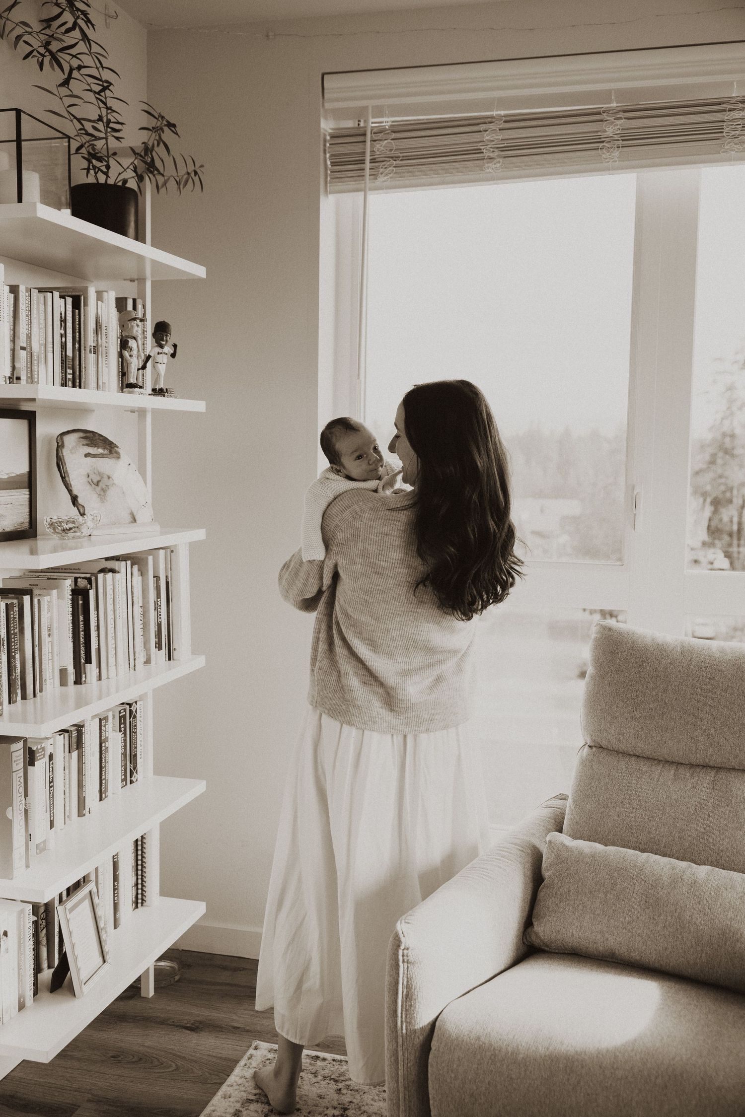 Mother holding newborn baby near sunlit window beside bookshelf in cozy nursery room, sepia tone.