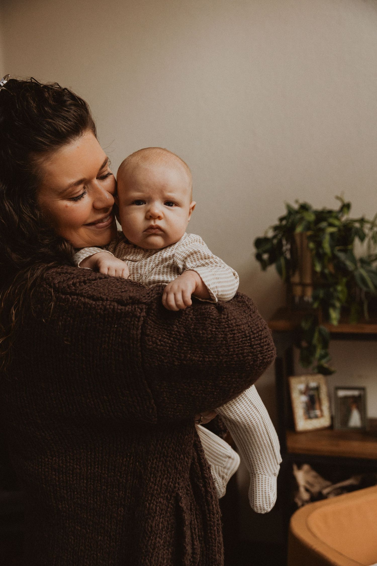Mother in brown knit sweater cuddling alert newborn baby against her cheek indoors.