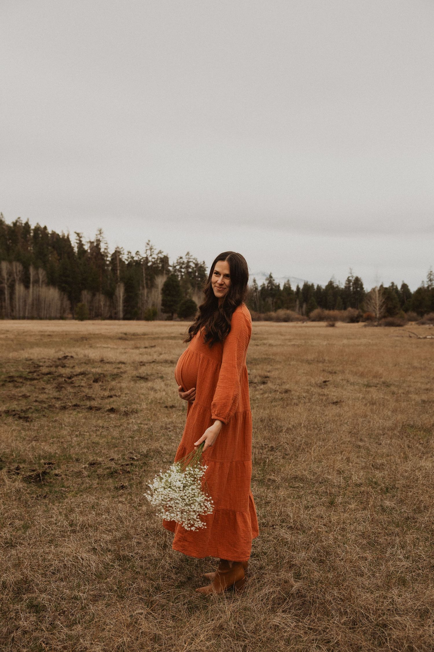 Smiling pregnant woman in rust orange maxi dress holding white wildflowers in an open field.
