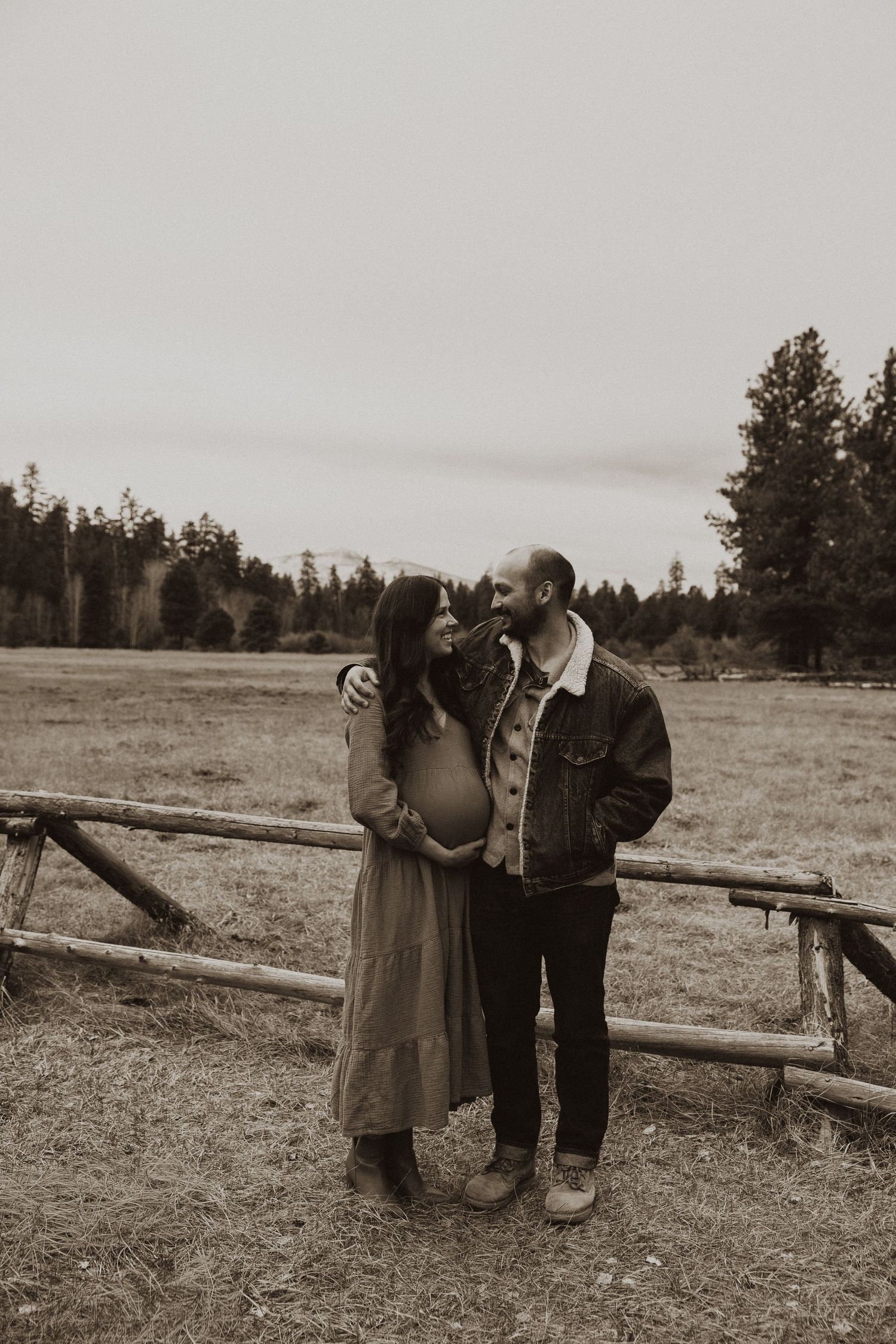 Expecting couple shares a tender moment by a rustic wooden fence in a sepia-toned outdoor maternity portrait.