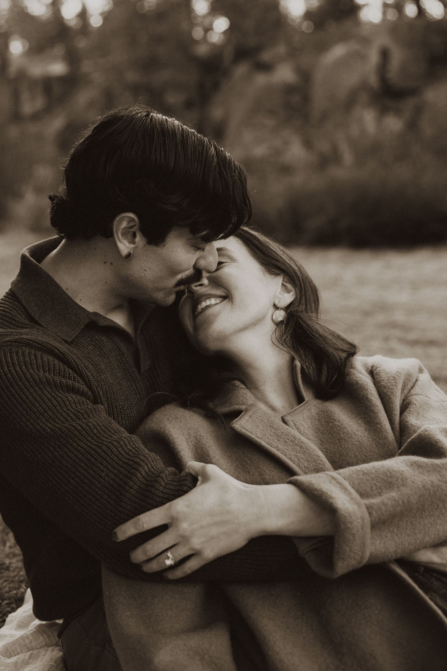 A couple shares a tender romantic moment outdoors, the man kissing her forehead as she smiles in sepia tones.