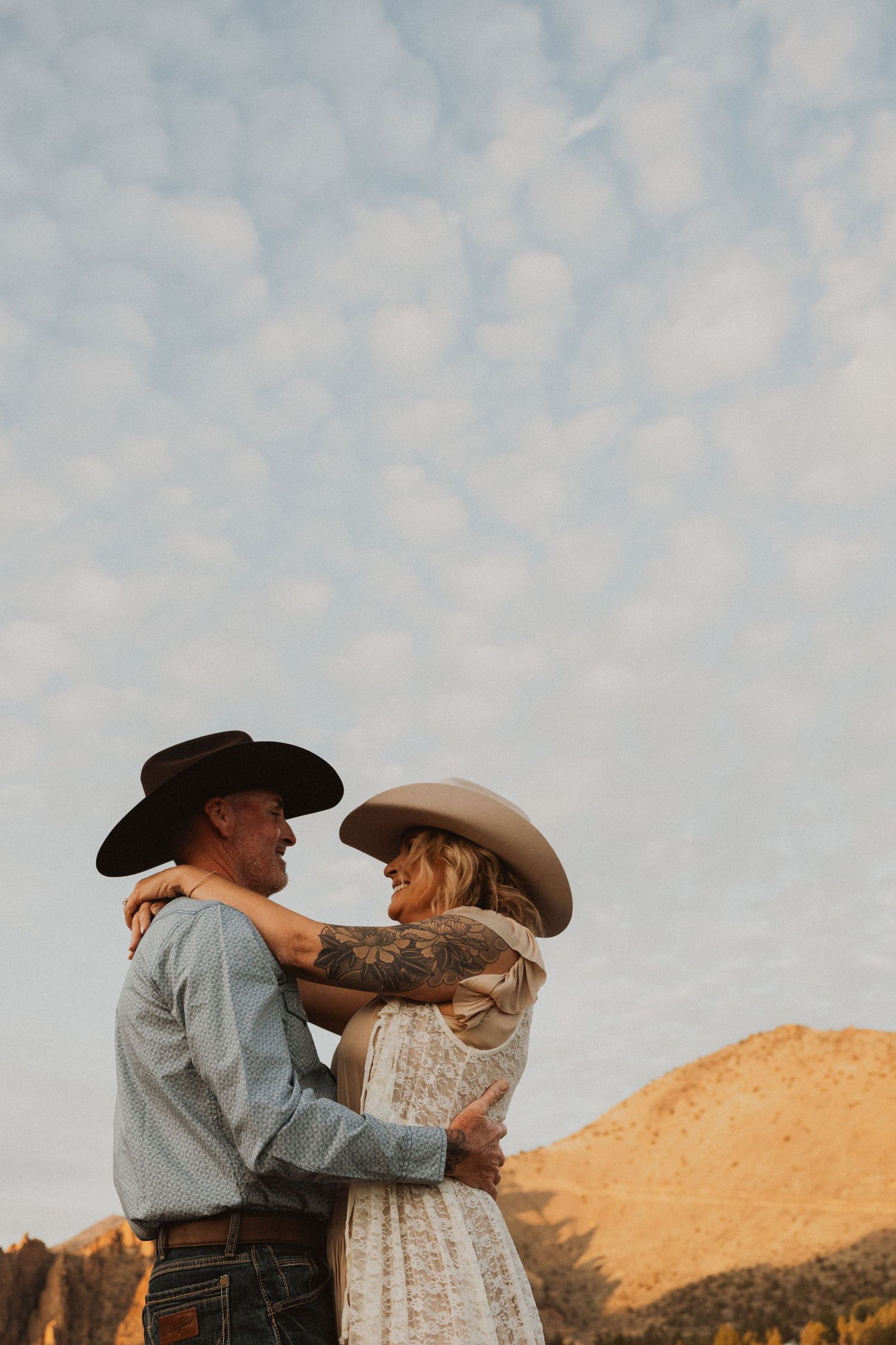 A couple in cowboy hats embracing outdoors with mountains and a cloudy sky backdrop in a romantic western-style photo.