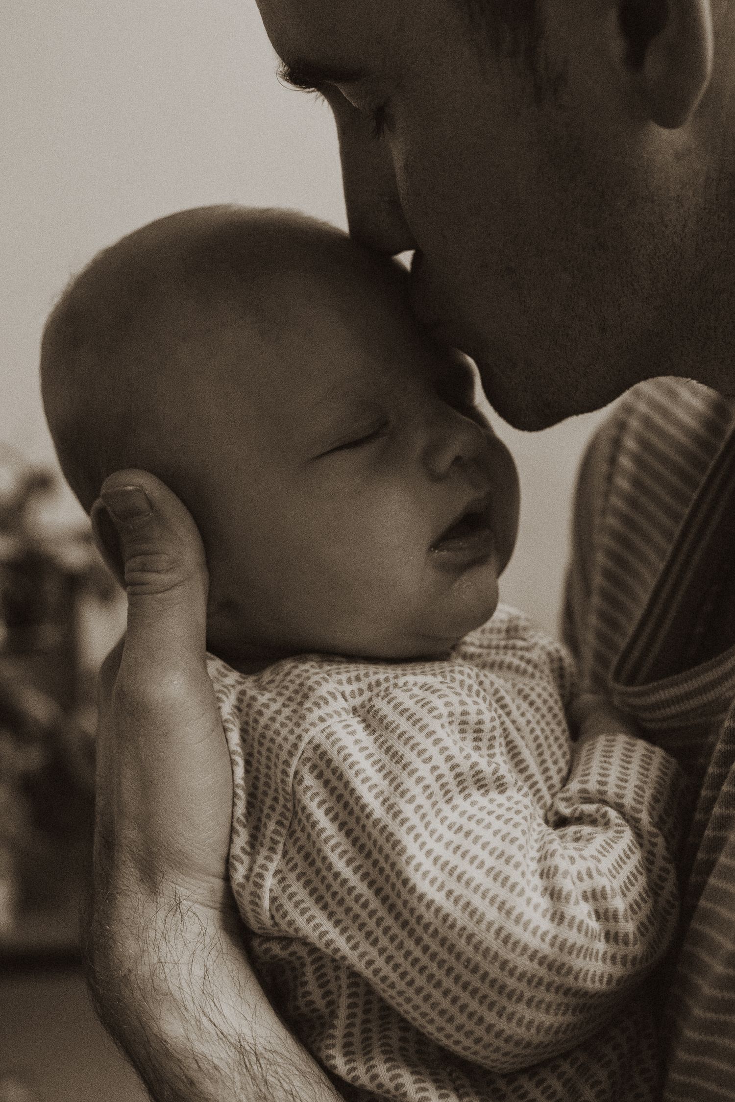 Father tenderly touching foreheads with his sleeping newborn baby in an intimate sepia portrait.