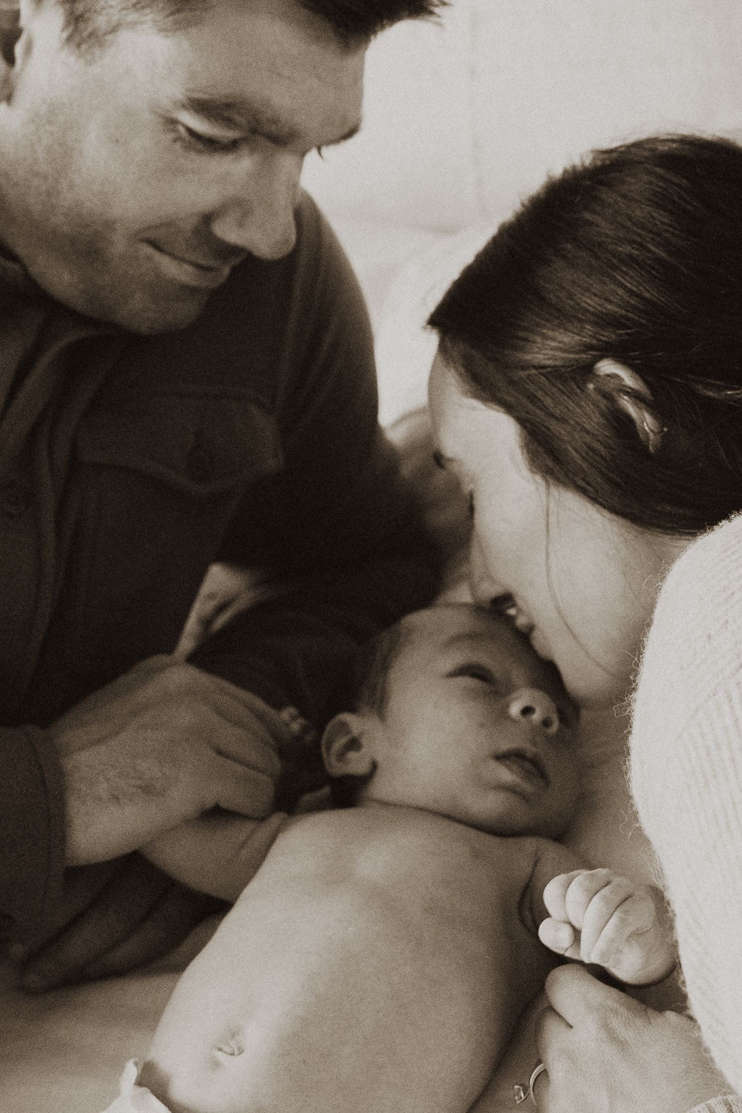 New parents lovingly gazing at their newborn baby together in a warm sepia-toned family portrait.