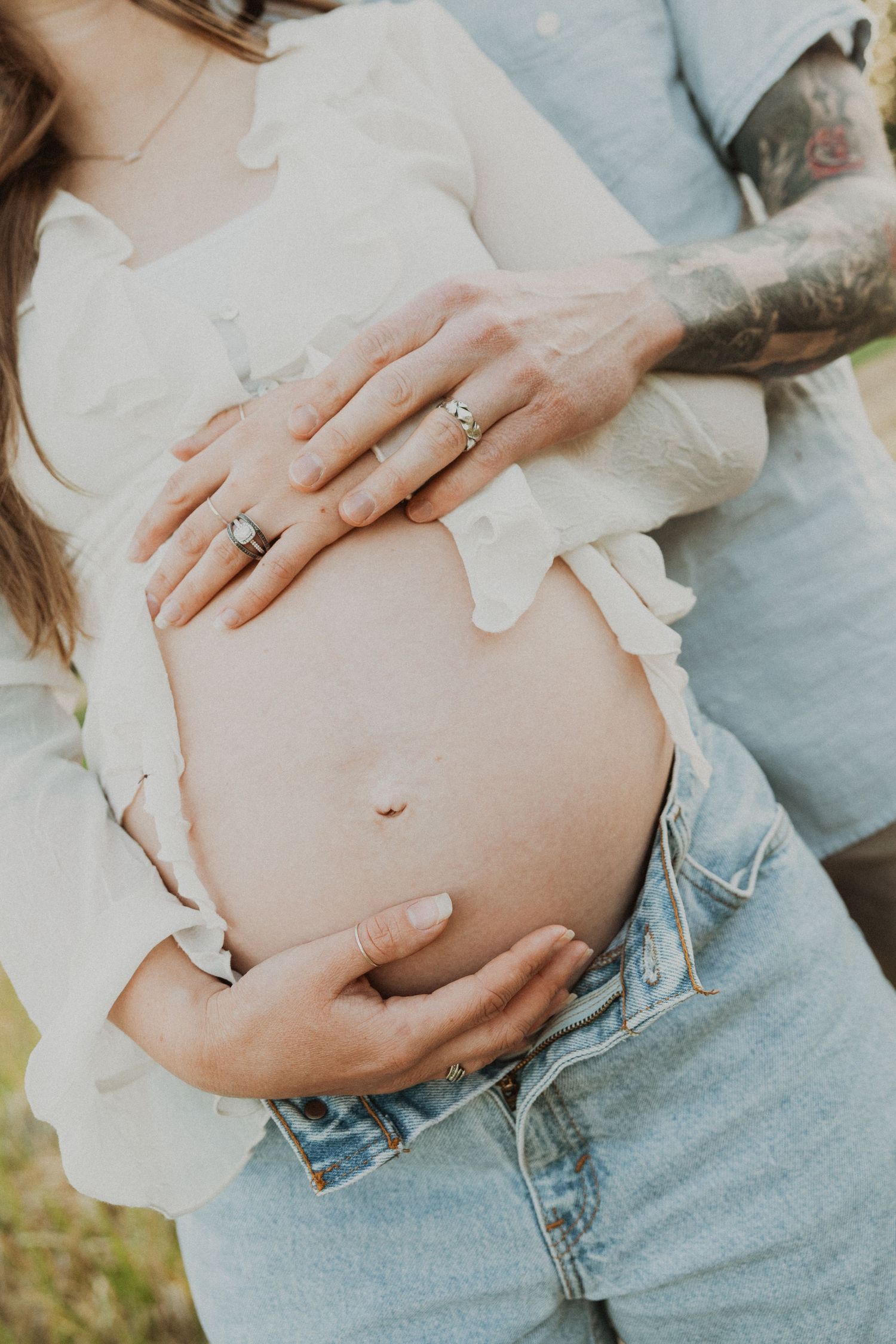 Close-up of couple cradling pregnant belly, tattooed partner embracing from behind outdoors.