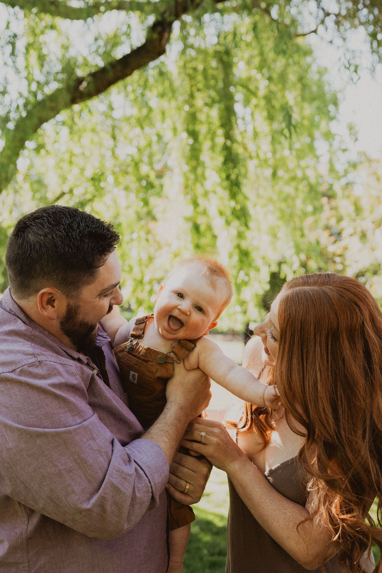 Happy redhead baby laughing while young parents hold him up outdoors under a lush green willow tree.