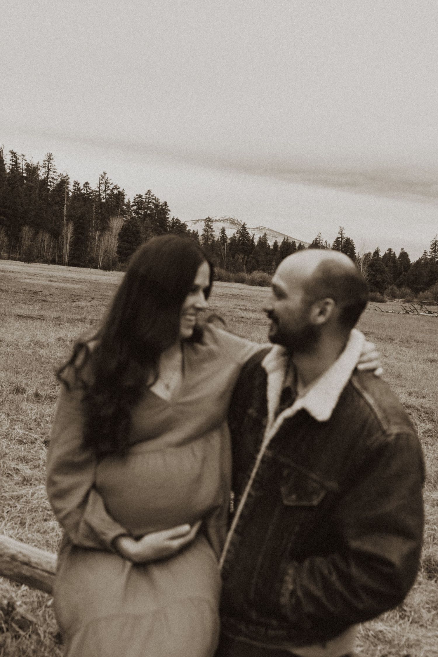 Couple shares an intimate moment in a sepia-toned maternity photo with pine trees and mountains in background.