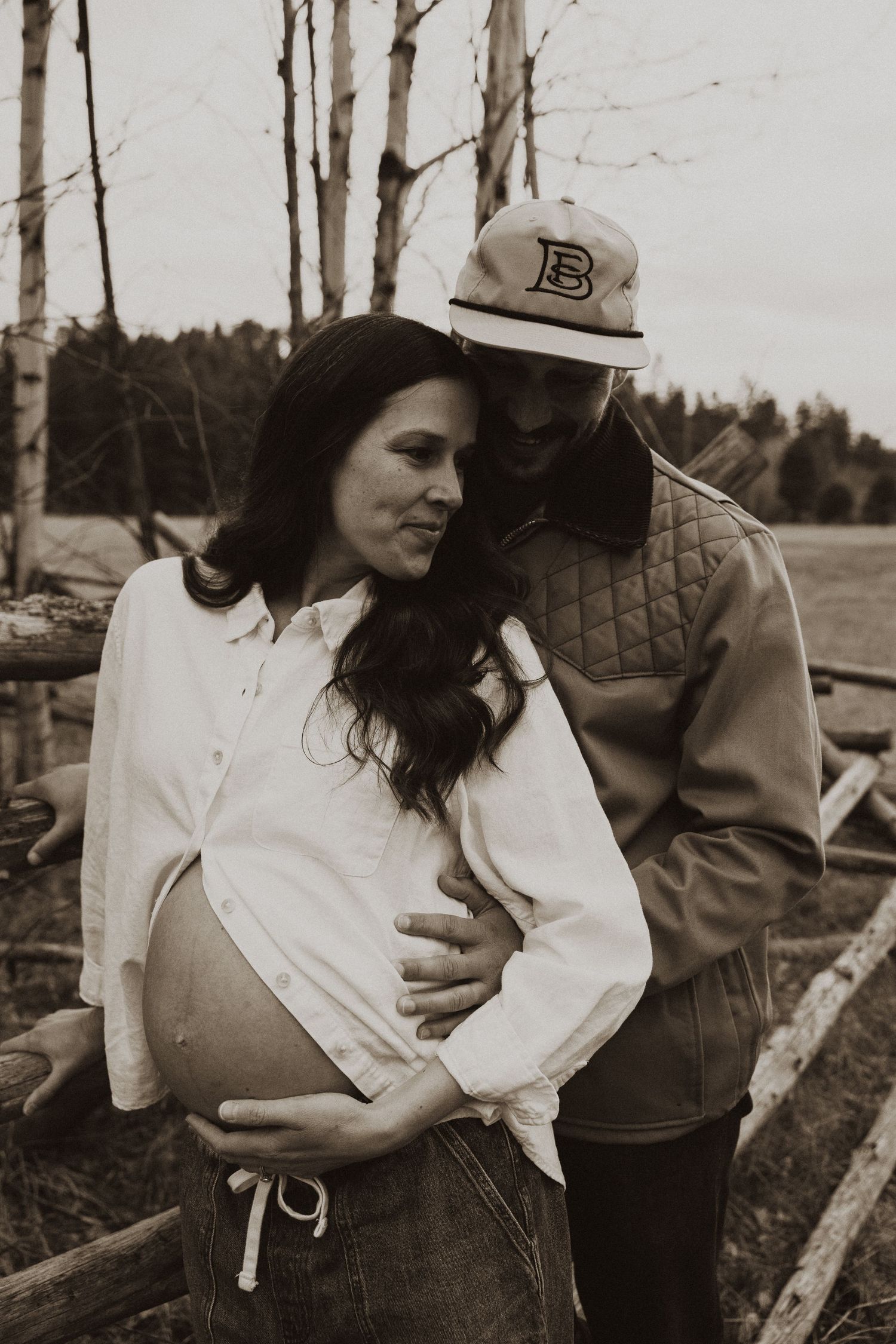 Sepia portrait of pregnant couple embracing near rustic wooden fence with birch trees behind them.