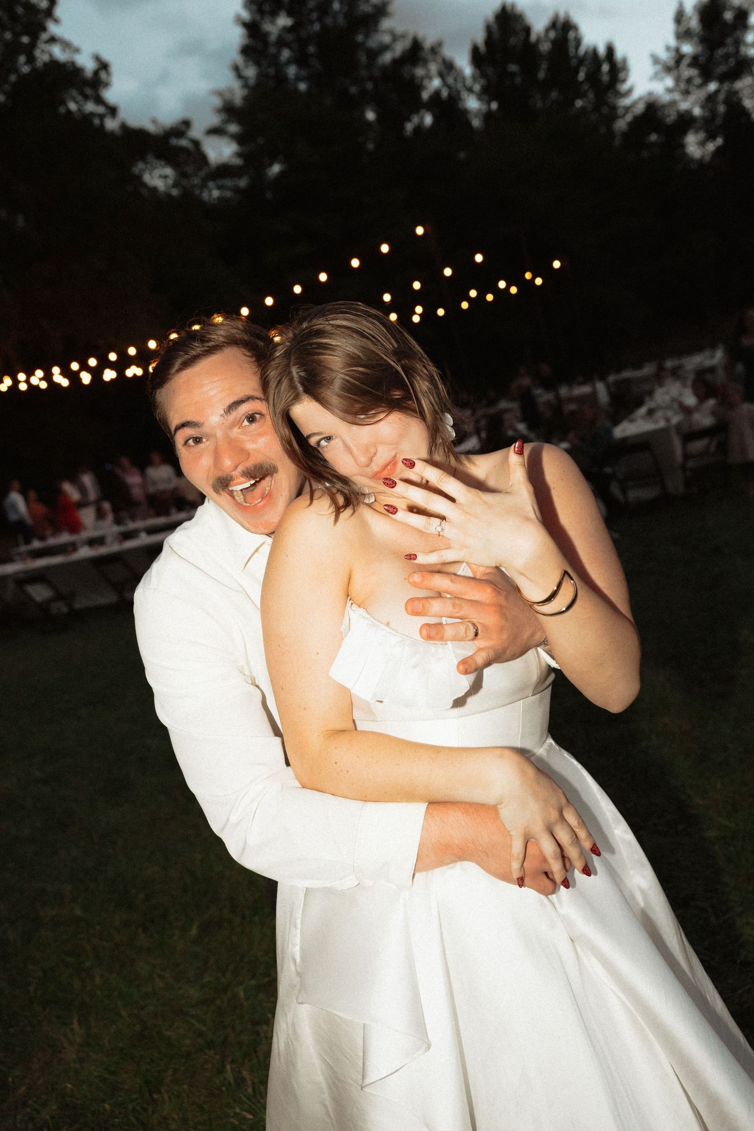 Joyful bride and groom embrace and laugh together outdoors under string lights during their evening wedding reception.