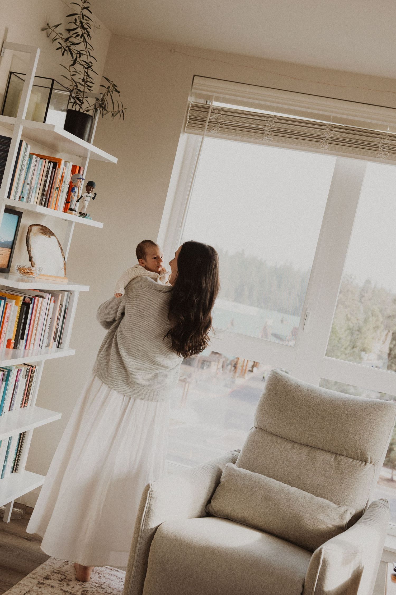 A mother holds her newborn baby near a large window beside a white bookshelf in a bright, modern nursery.
