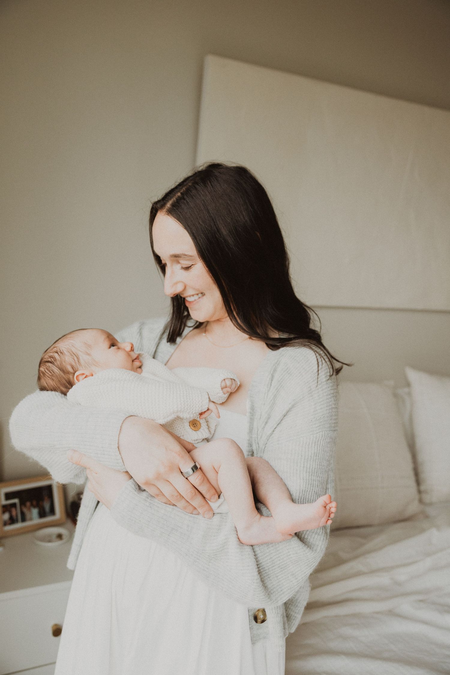 A smiling dark-haired mother in a grey sweater tenderly holds her newborn baby in a bright bedroom.