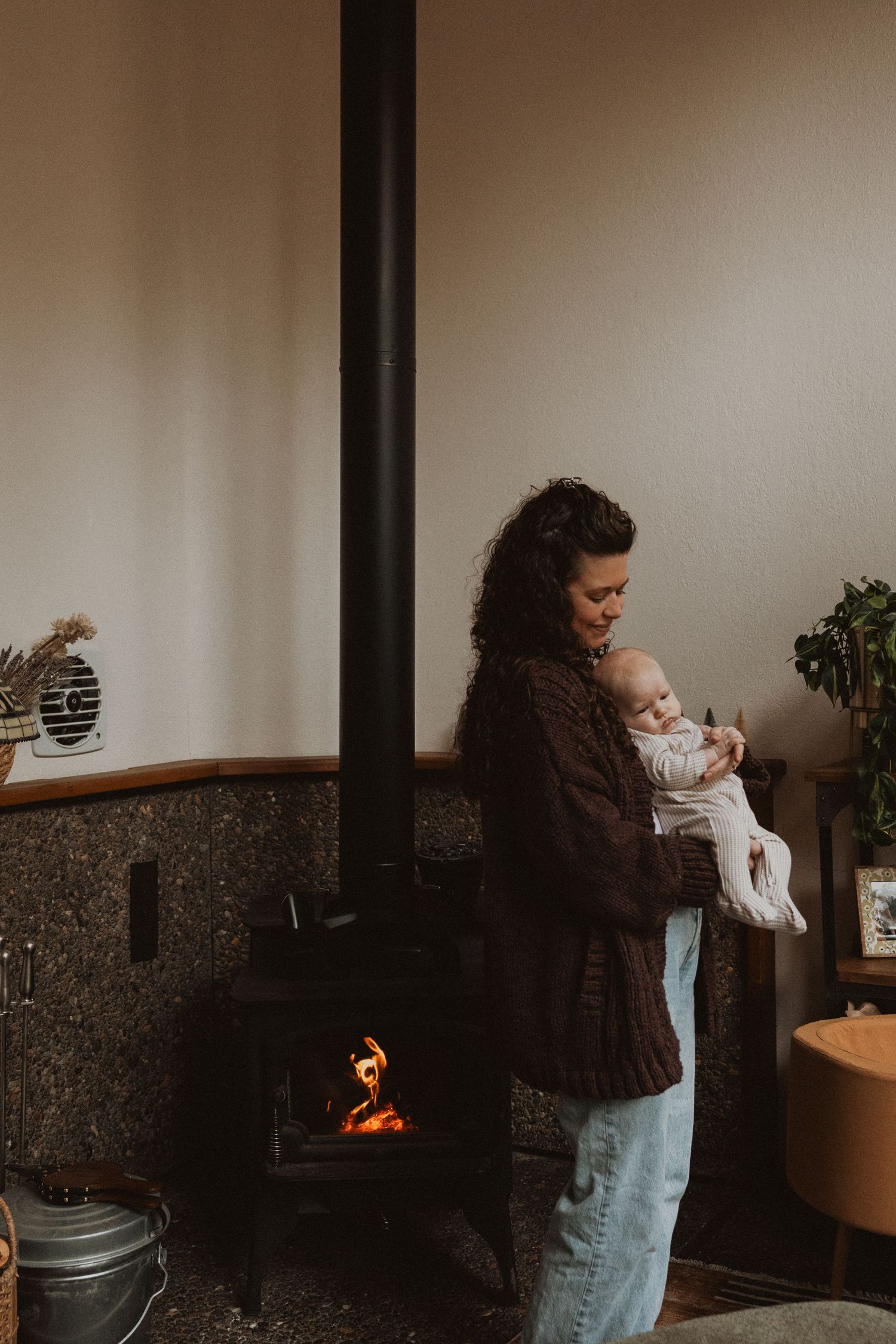 Mother in brown sweater holding newborn baby near a warm wood-burning fireplace at home.