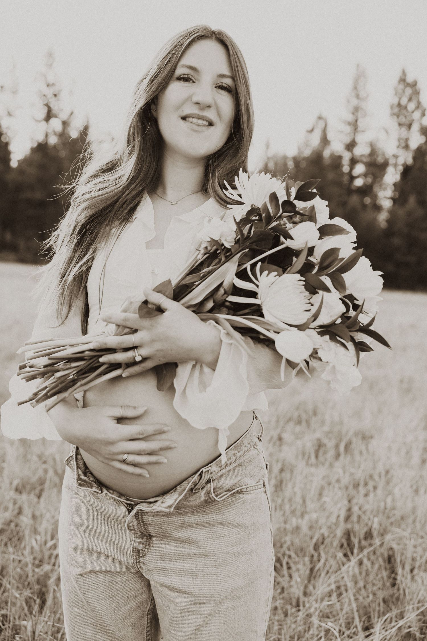 Smiling pregnant woman holding white floral bouquet in a field, sepia-toned maternity portrait.