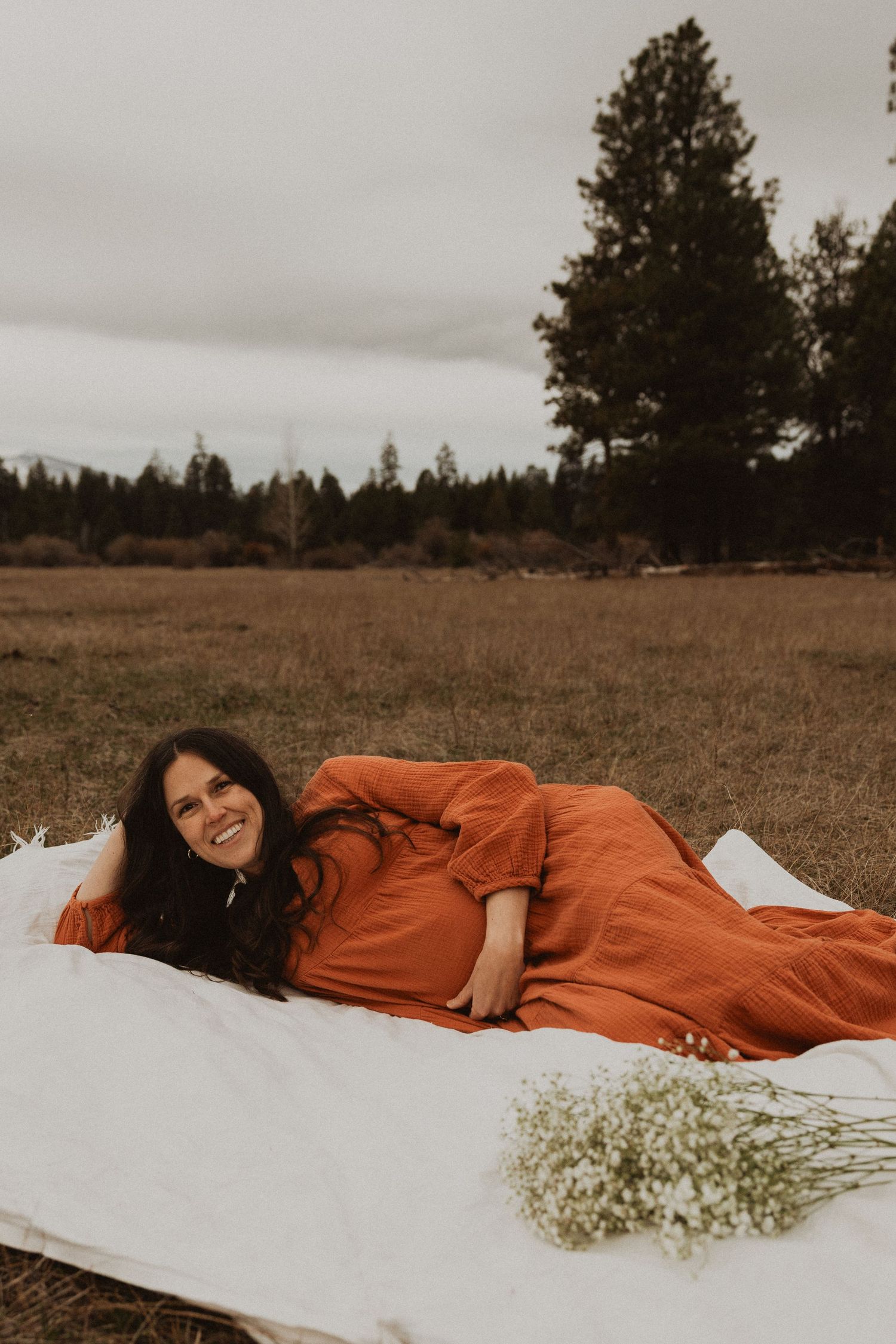 Smiling pregnant woman in rust orange maxi dress lying on white blanket in field with baby's breath bouquet.