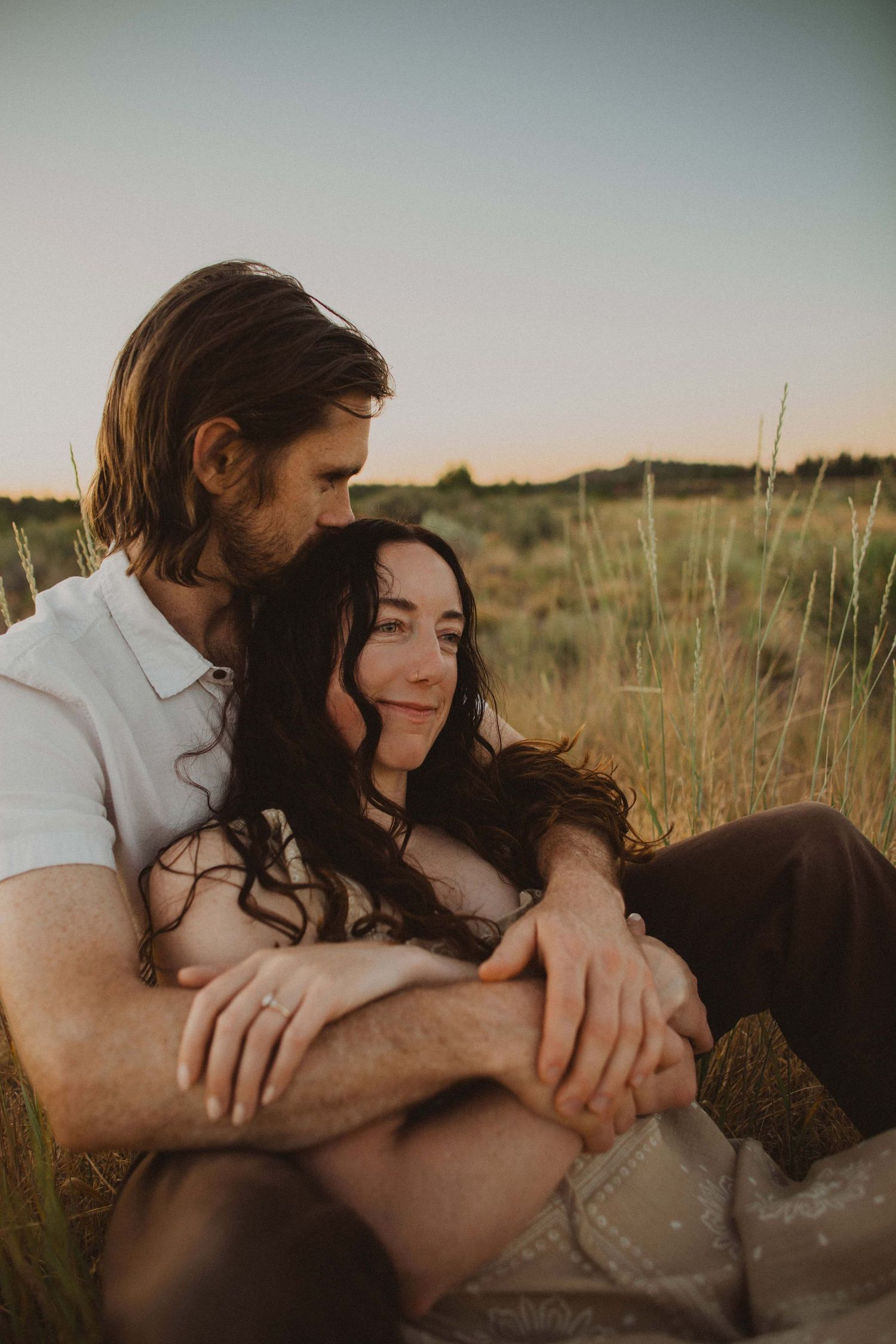 Two people share intimate moments while sitting in desert brush at sunset.