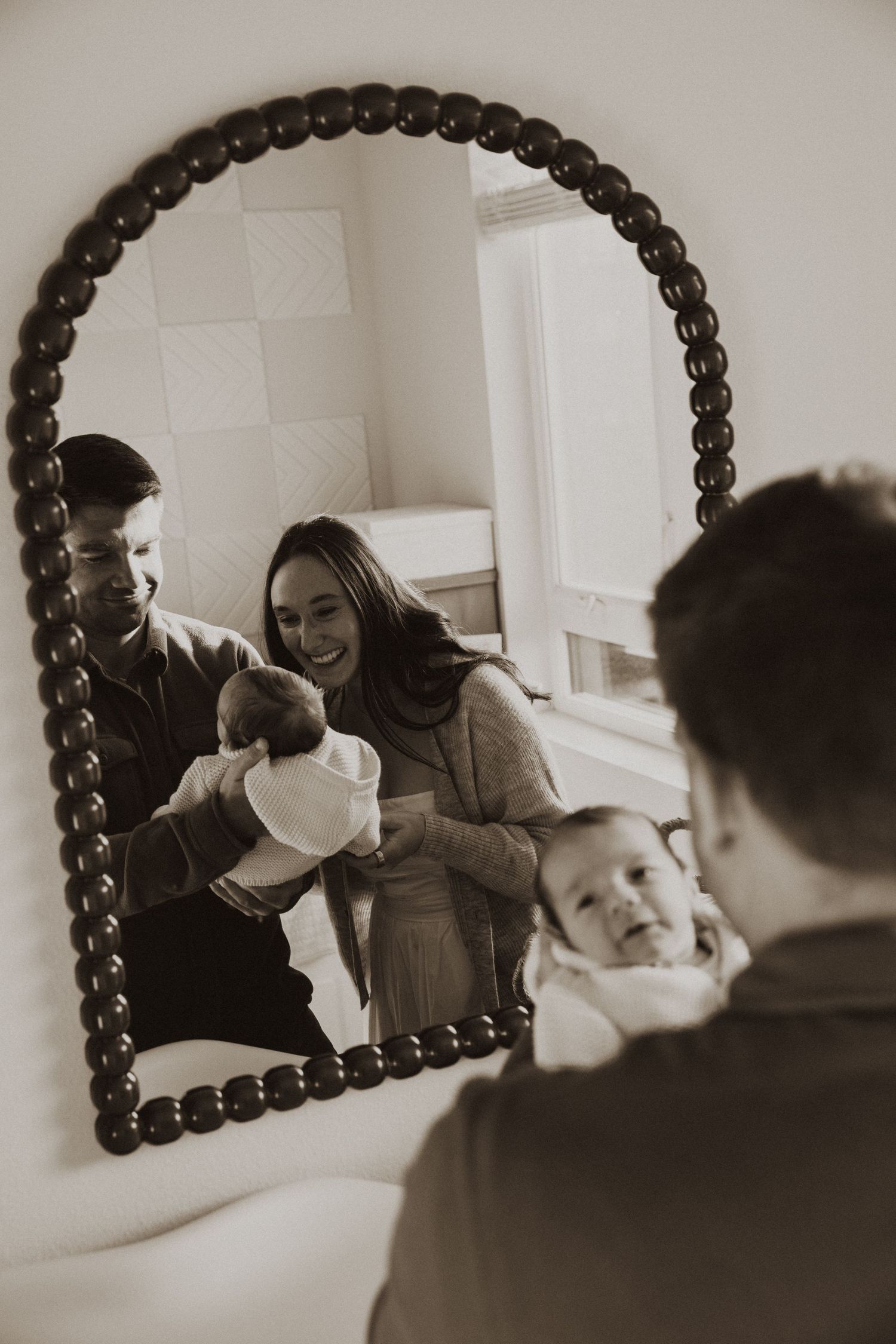 Parents smile at their newborn baby reflected in an arched beaded mirror in a black and white family photo.