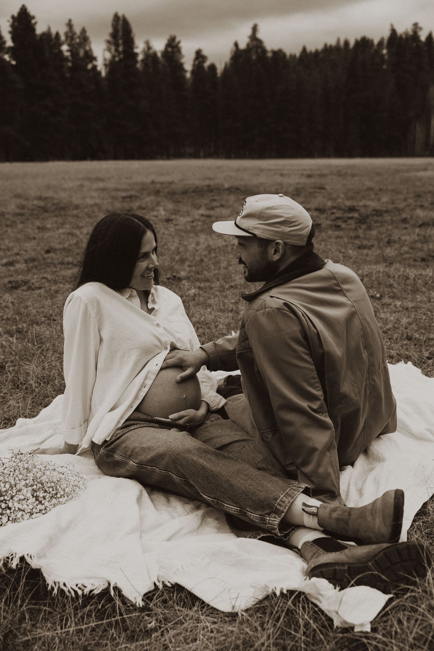 Black and white maternity photo of couple sitting on blanket in field, man touching pregnant belly.