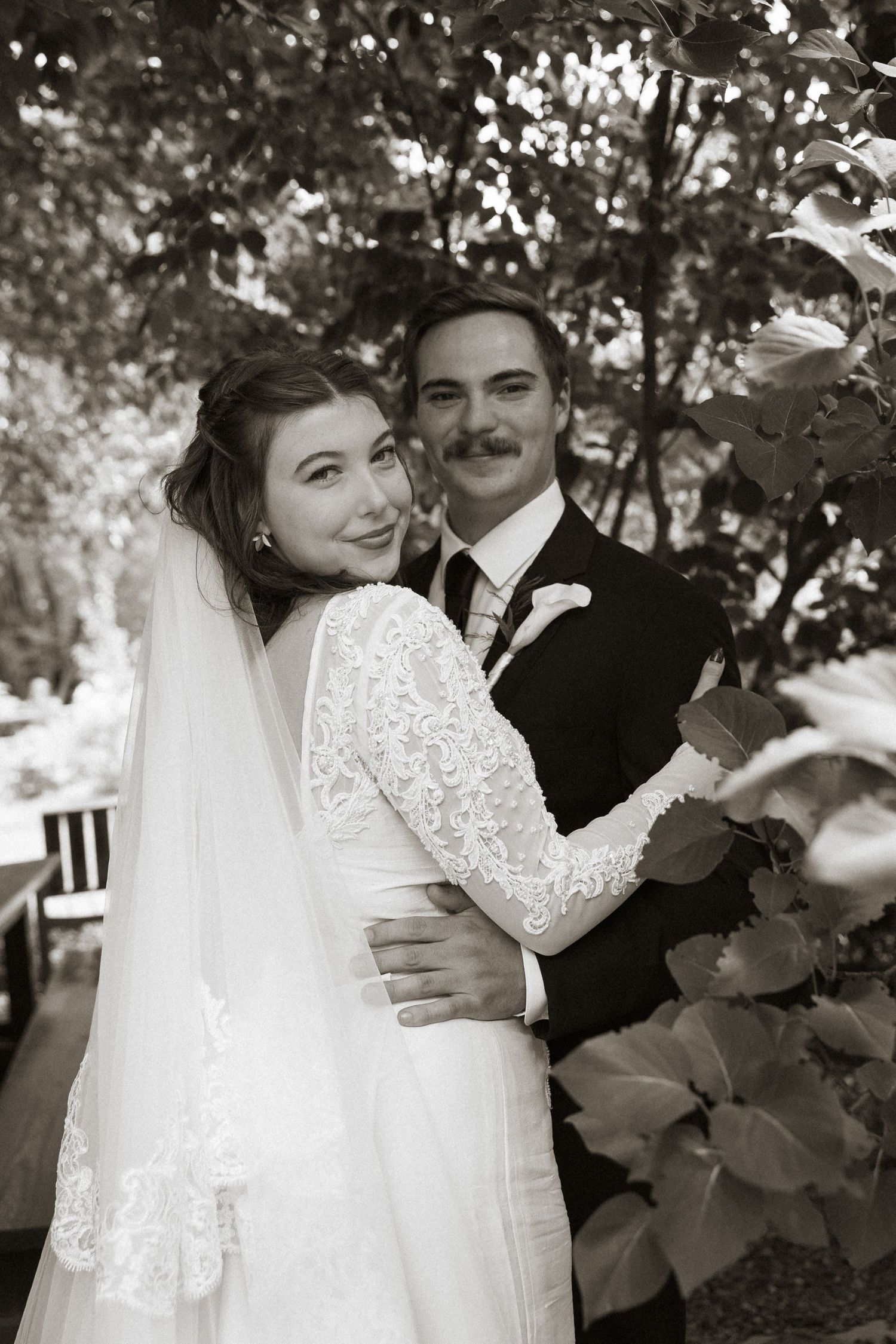 Black and white photo of a smiling bride in lace gown and veil embracing a groom with mustache outdoors.