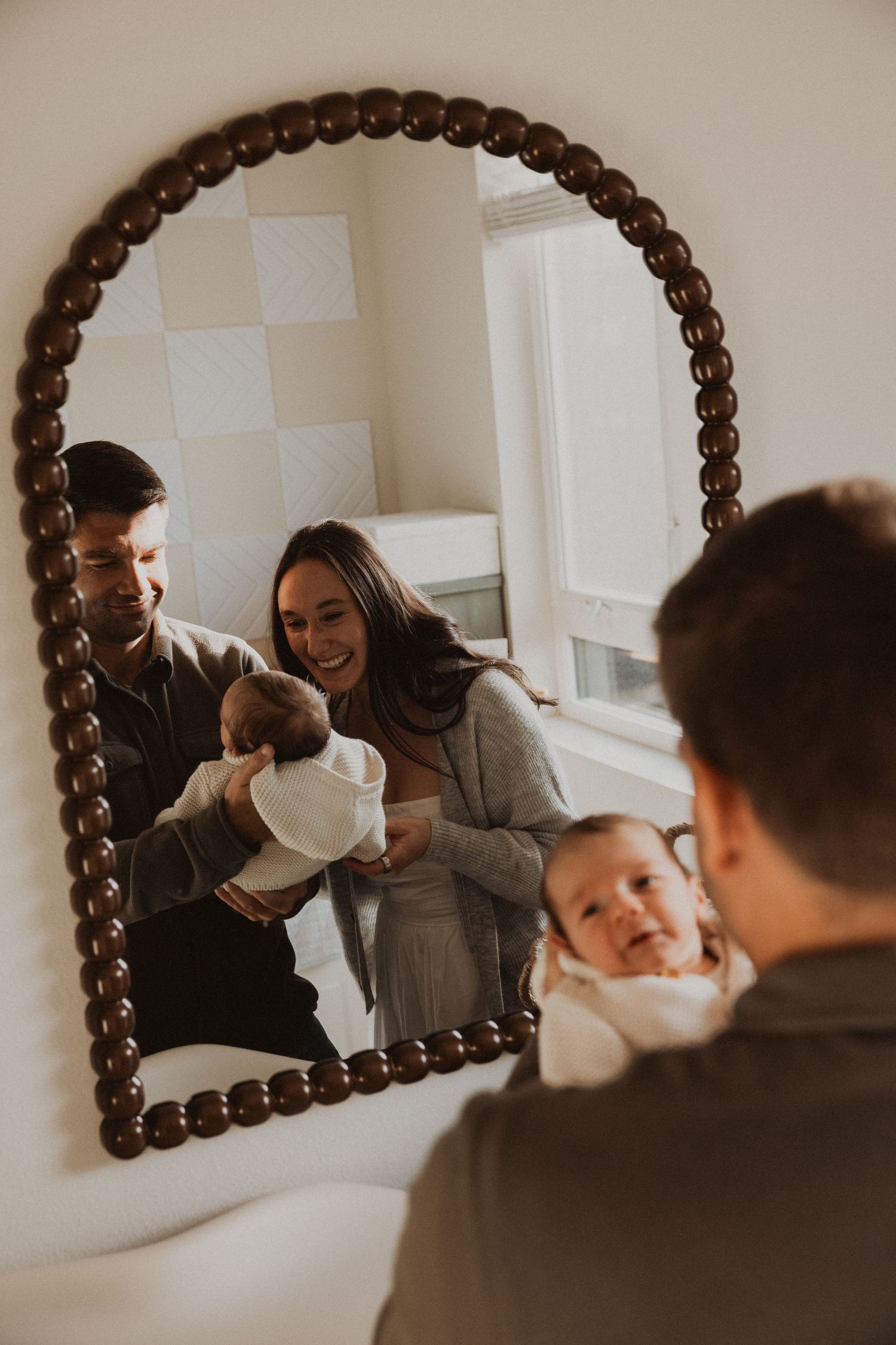 Family with newborn baby reflected in arched beaded mirror in a cozy nursery.