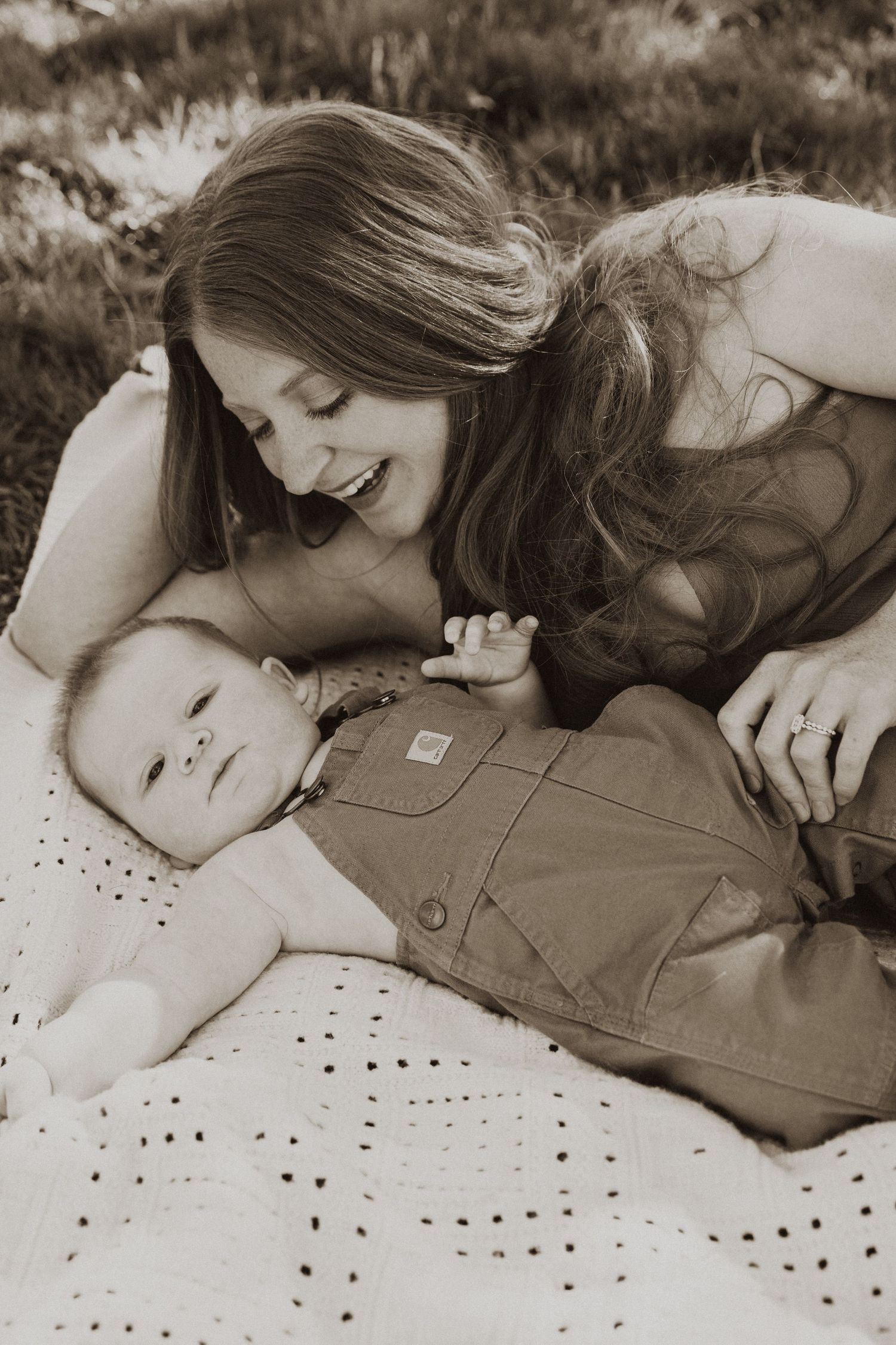 Smiling mother lying on a blanket outdoors, lovingly gazing at her baby in overalls in a sepia-toned photo.