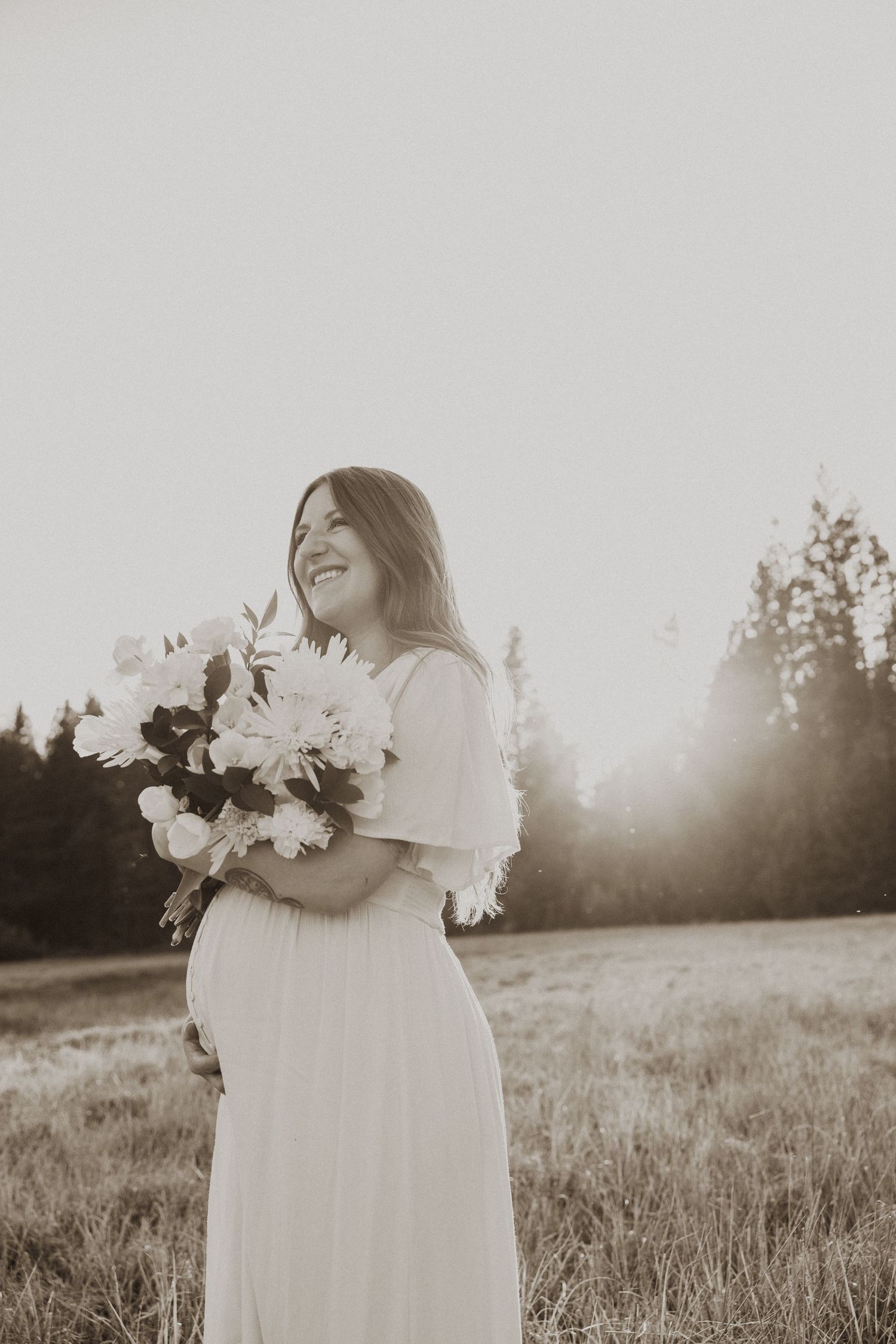 Smiling pregnant woman in white dress holding bouquet of white flowers in sunlit field.