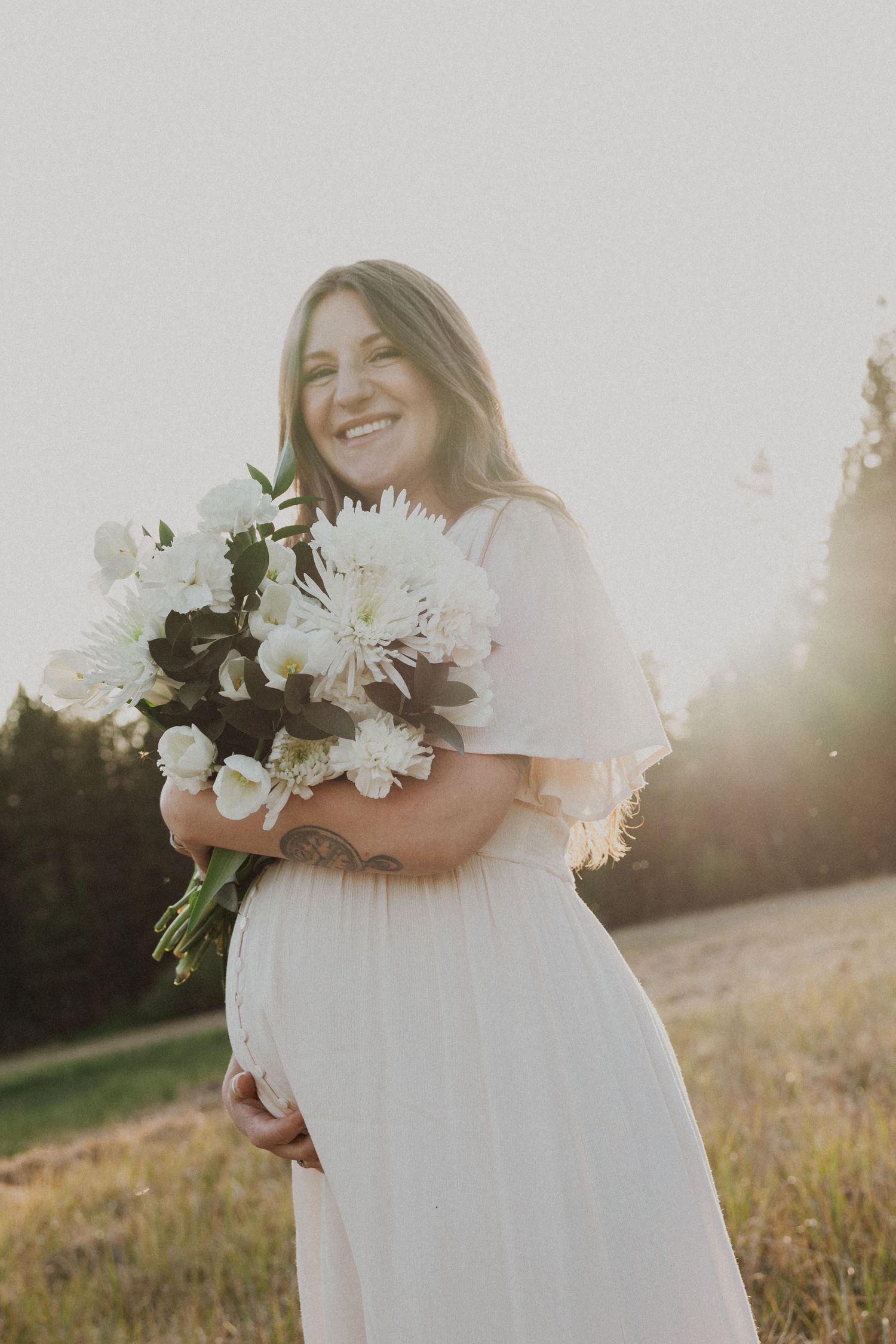 Smiling pregnant woman in white dress holding large white flower bouquet outdoors in golden sunlight.