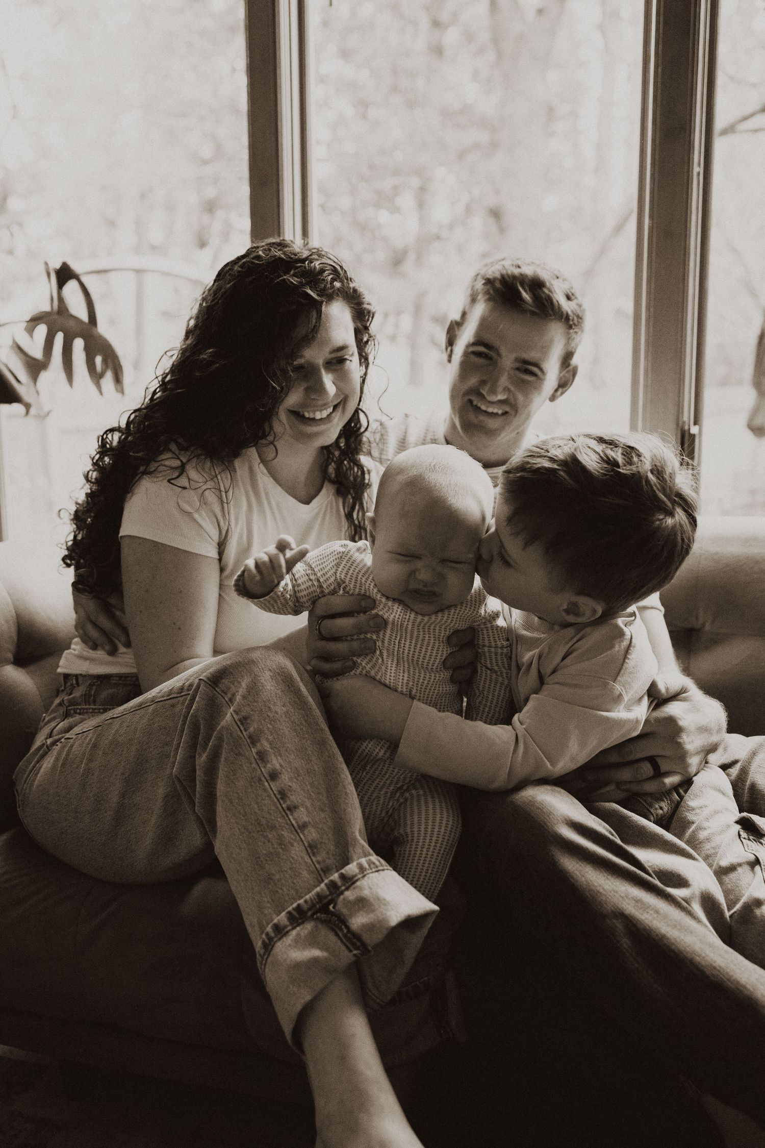 Joyful family of four with newborn baby sharing tender moment on couch in sepia tone.