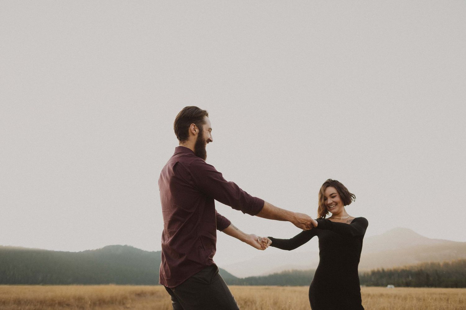 Two people dance playfully in a scenic field during golden hour with mountains in the distance.