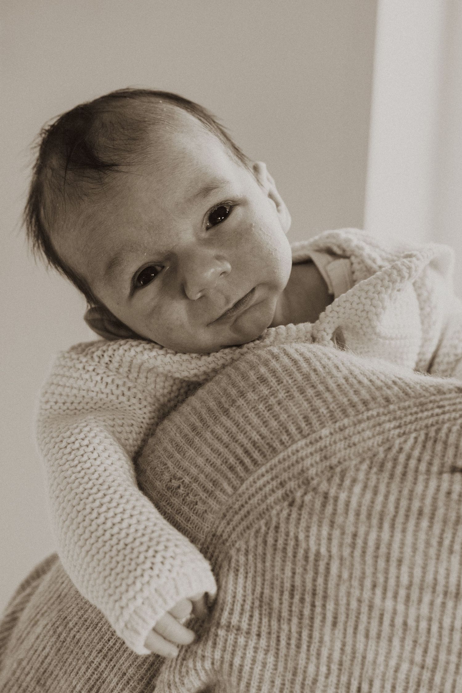 Newborn baby with dark hair resting on a knitted blanket, looking up in a soft sepia-toned portrait photo.
