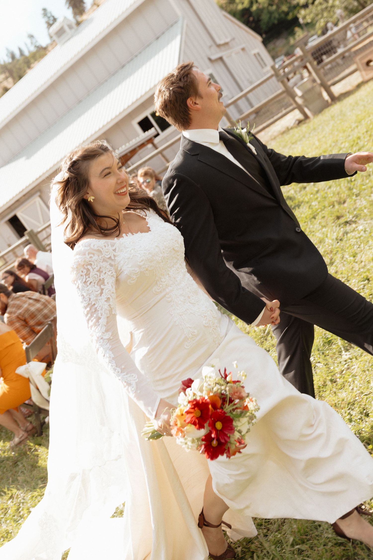 Joyful bride and groom walk hand in hand after outdoor wedding ceremony, bride holding red floral bouquet.