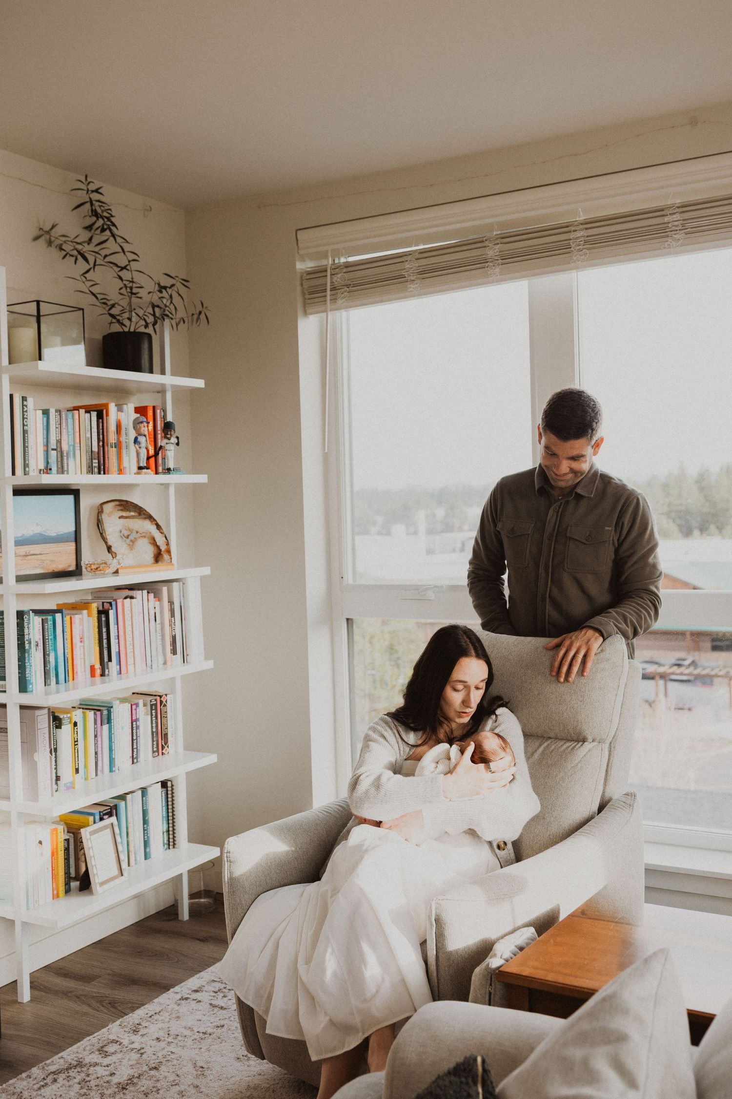 A couple admires their newborn baby in a cozy living room with a bookshelf, the mother seated in a gray armchair.