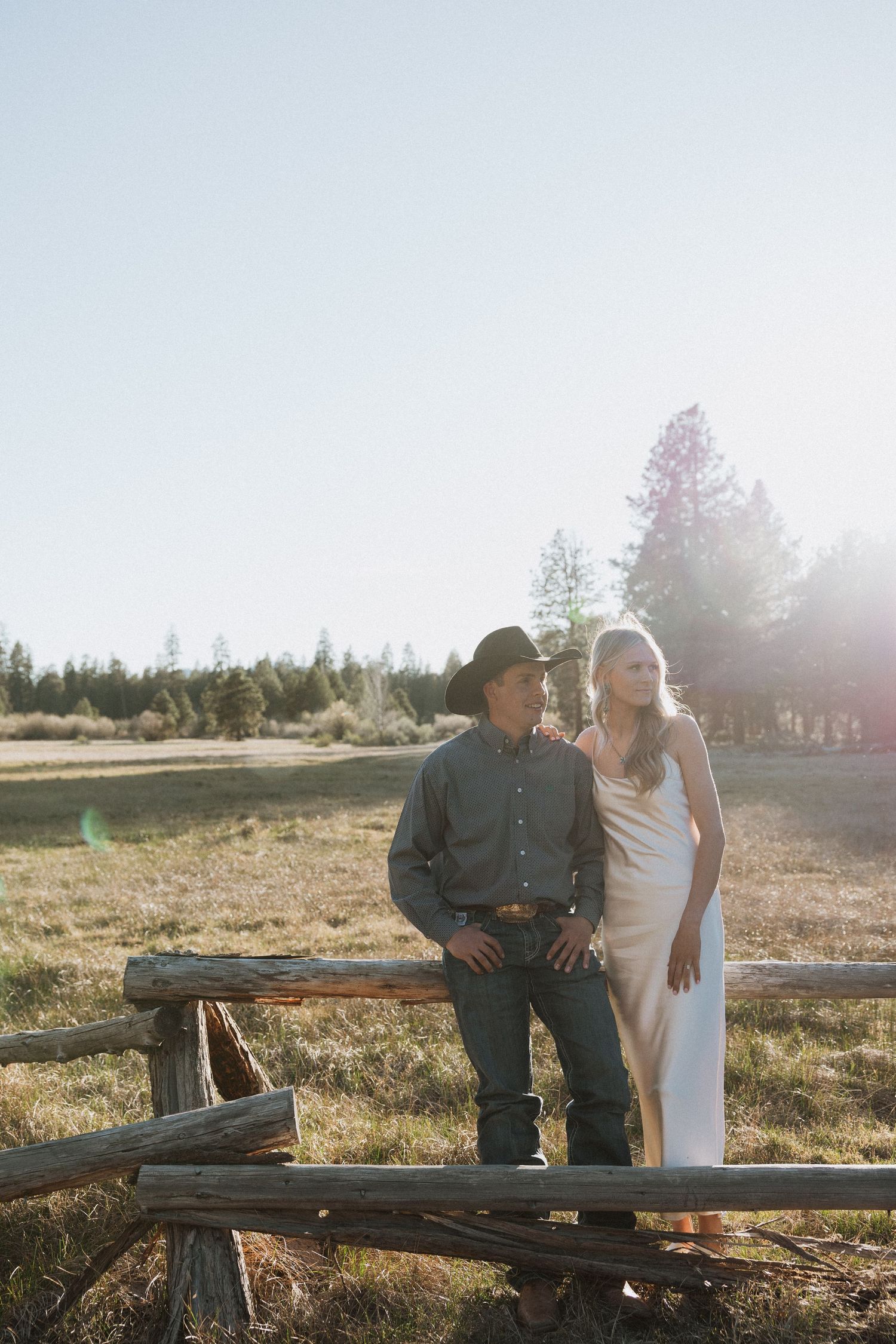 Couple posing by rustic wooden fence in sunny meadow, man in cowboy hat and woman in white slip dress.
