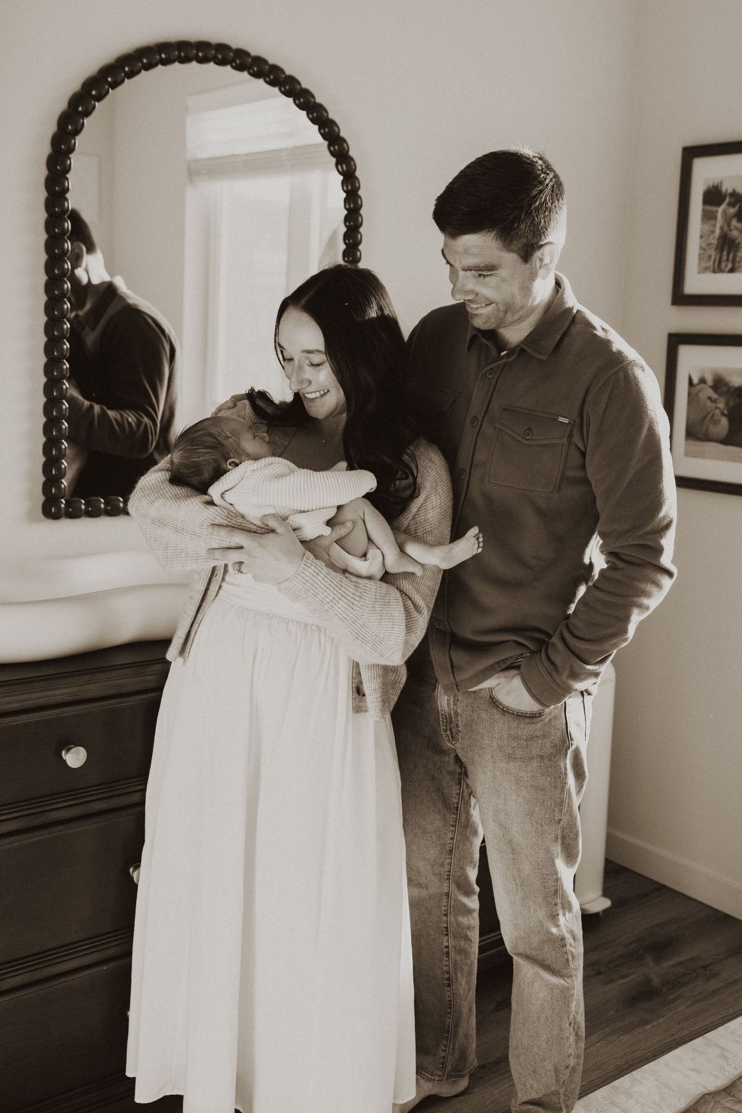 Black and white photo of new parents smiling at their newborn baby in a cozy bedroom with a mirror.