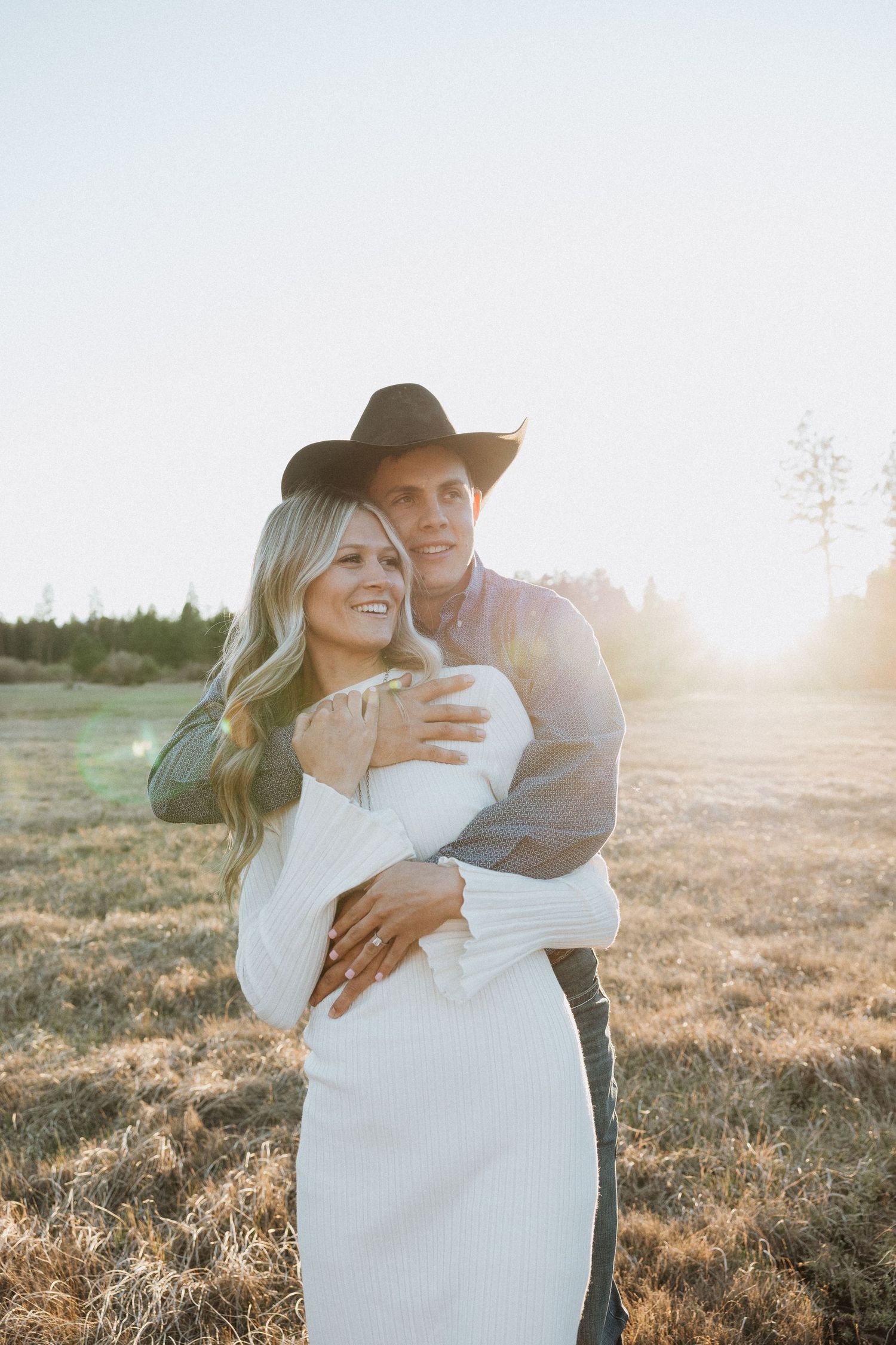 A couple embraces in a golden sunlit field, man in a black cowboy hat hugging a smiling blonde woman in a white dress.
