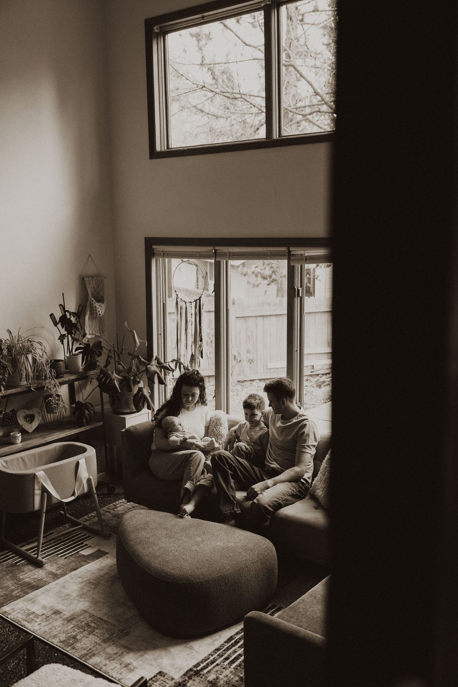 Sepia-toned photo of family with newborn sitting on couch in cozy plant-filled living room.