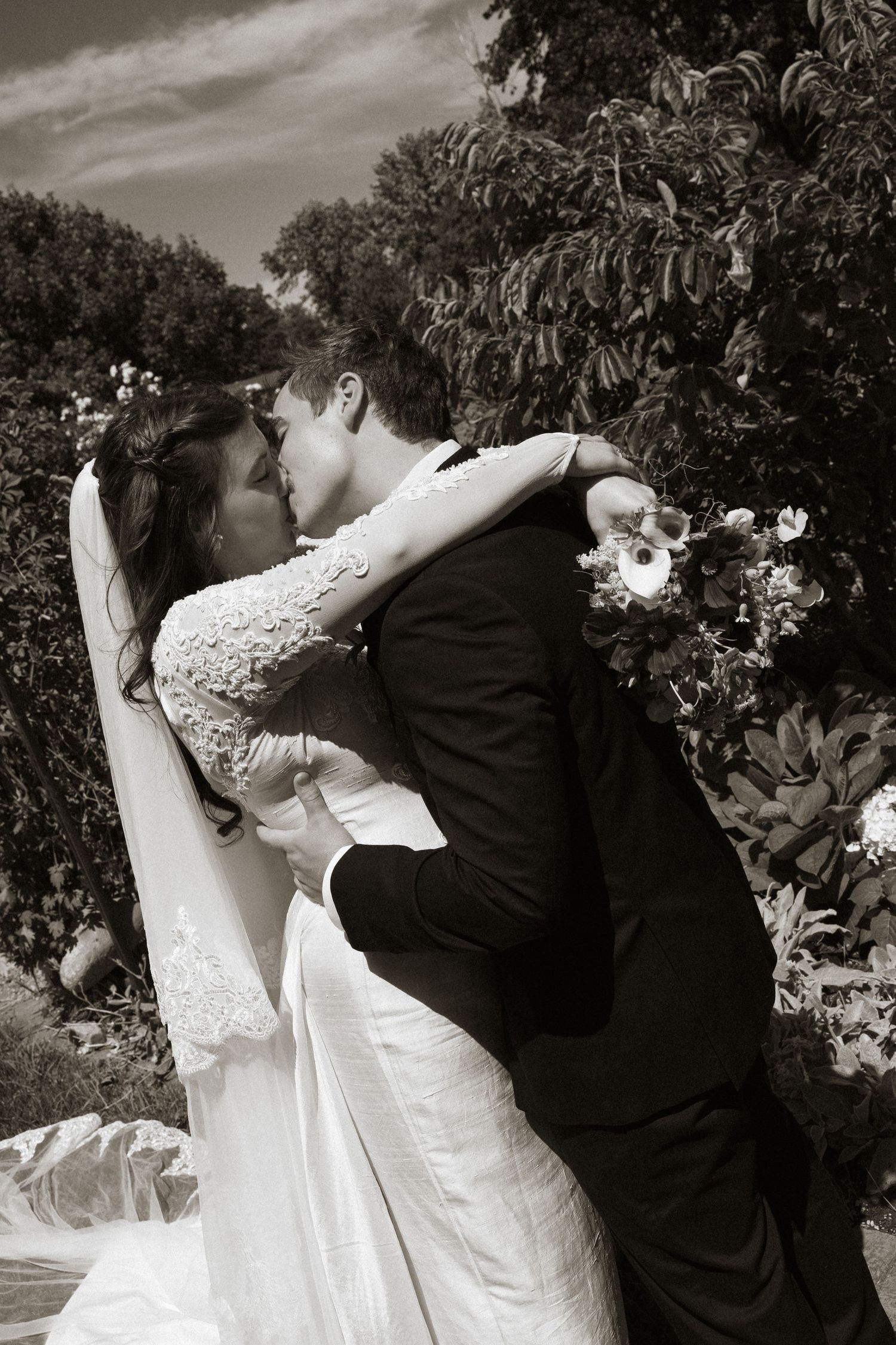 Bride and groom share a romantic kiss outdoors, surrounded by lush greenery, in a classic black and white wedding photo.
