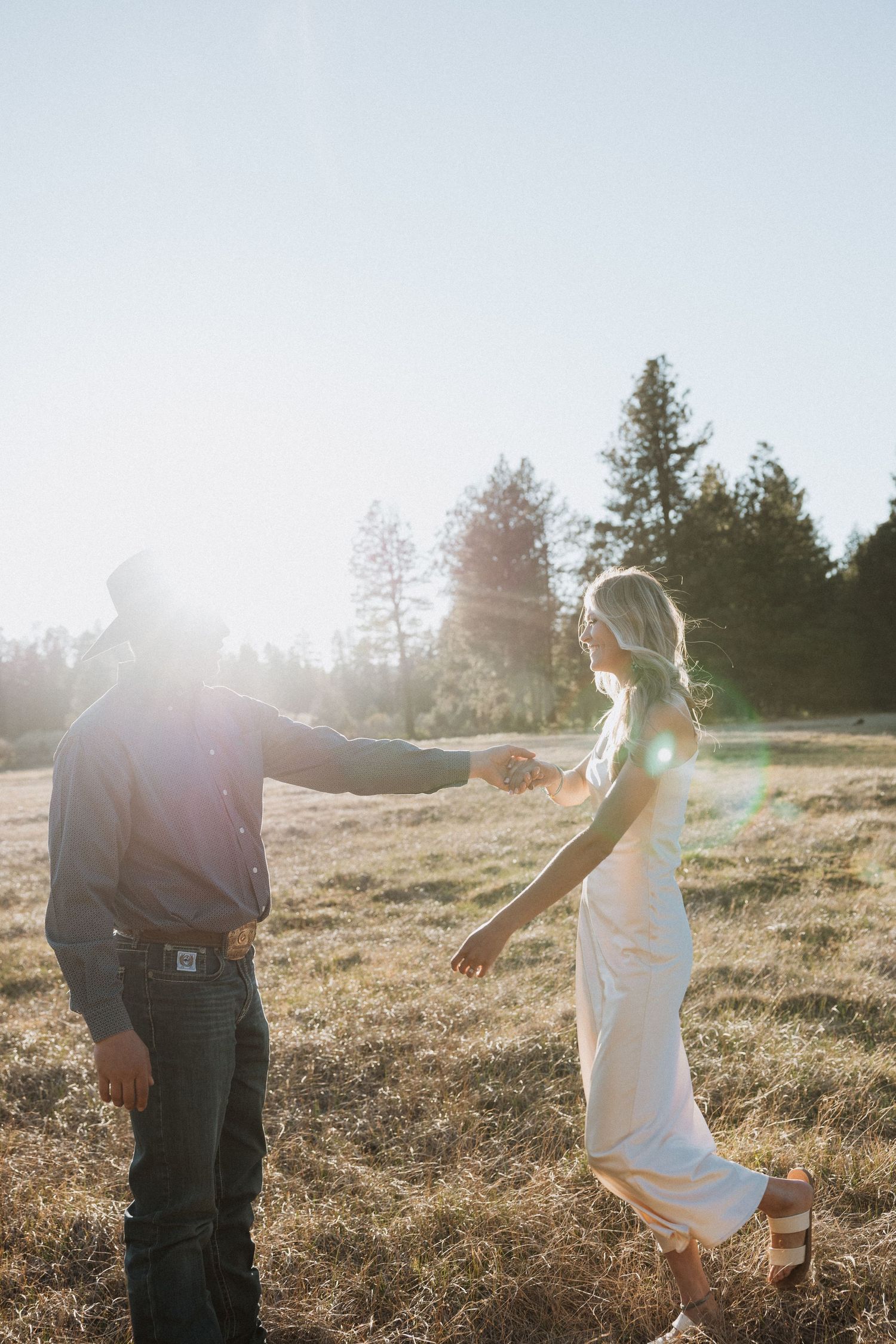 Couple holding hands in a sunlit field with evergreen trees, woman in white dress, man in jeans and hat.