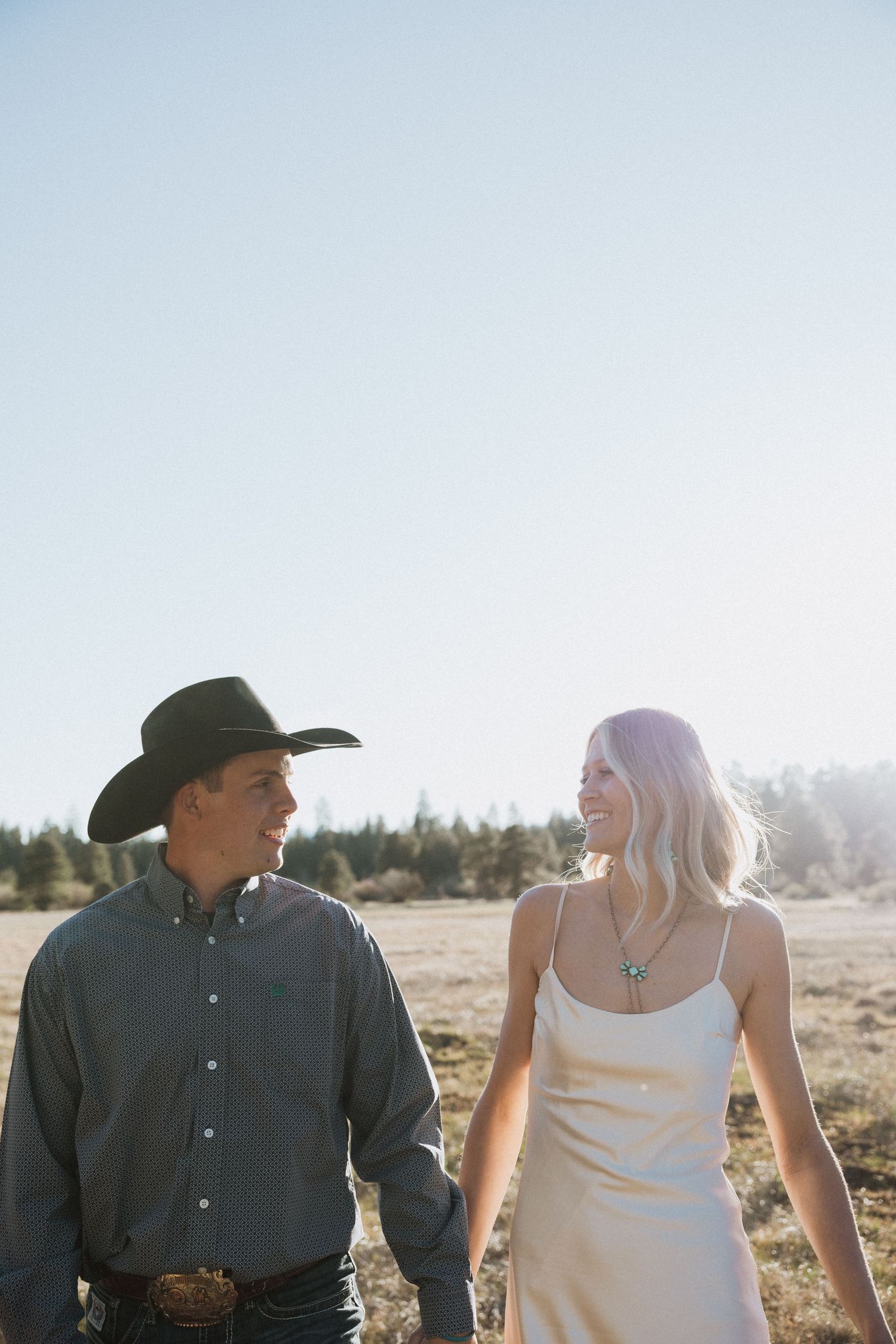 A smiling couple walks through a sunlit field, man wearing a black cowboy hat and denim shirt, woman in a white slip dress.
