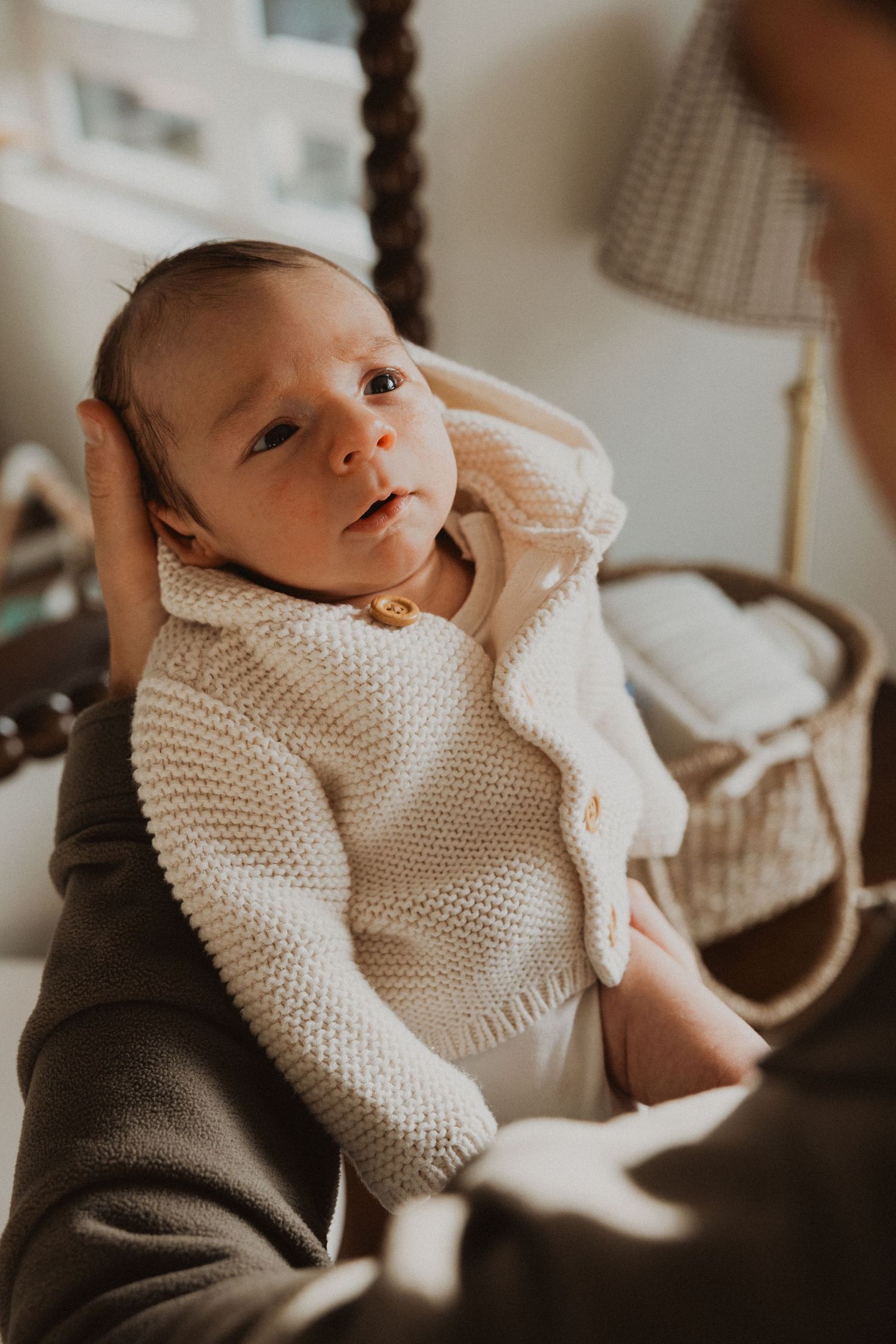 Newborn baby in a cream knit sweater being held by a parent near a wicker bassinet in a softly lit nursery.