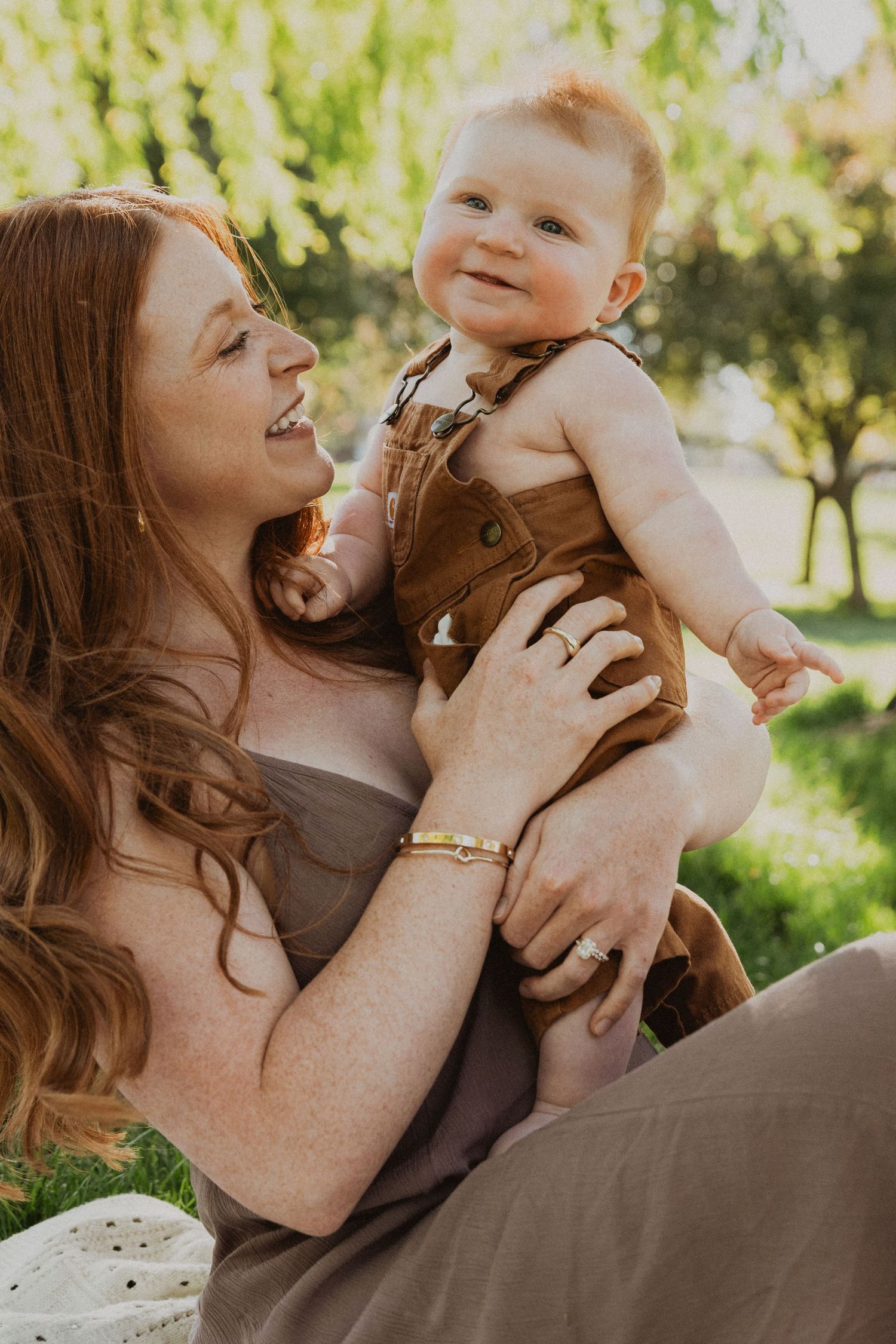 A redheaded mother smiles while holding her happy baby in brown overalls outdoors in a sunny green park.