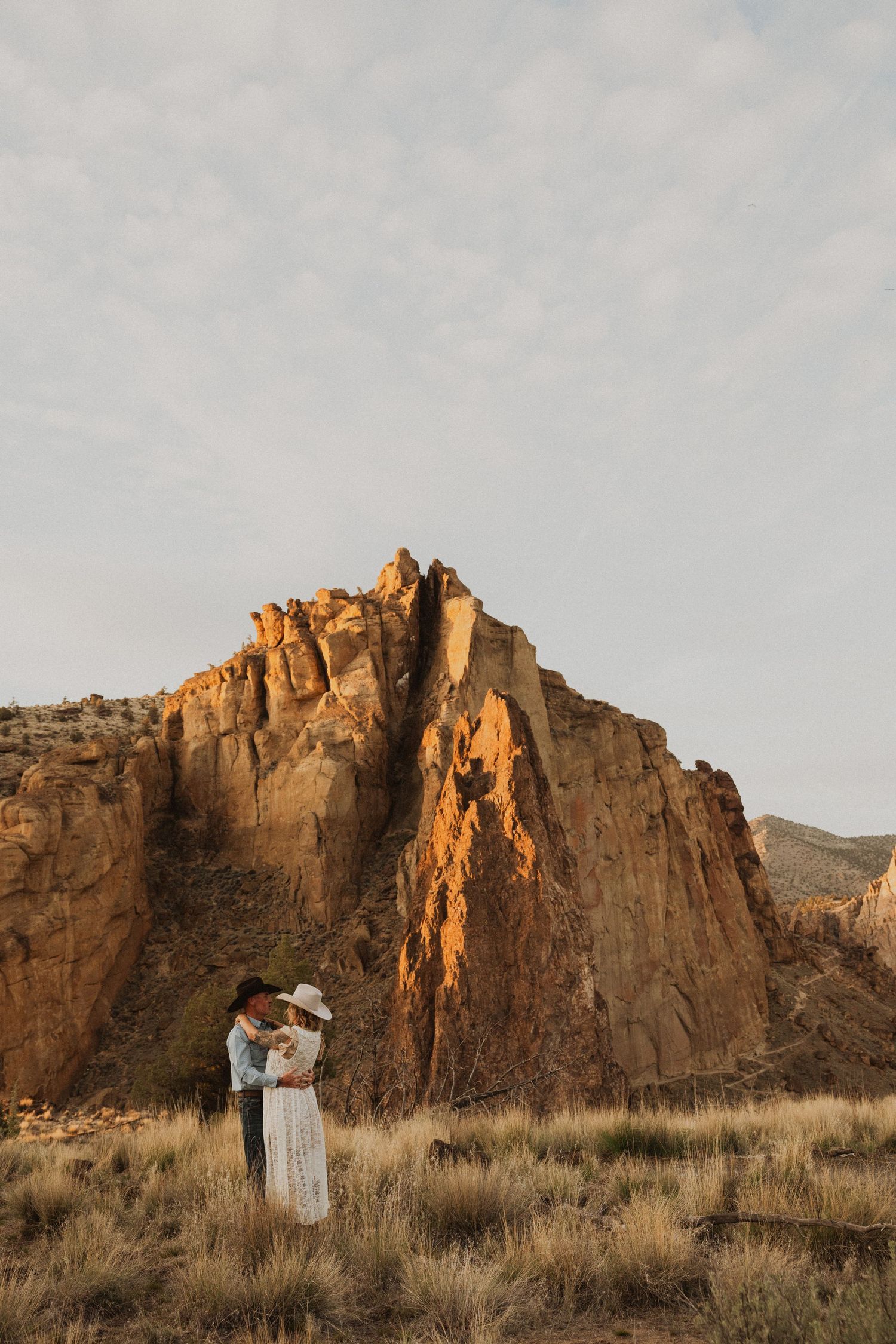 Couple embracing in front of dramatic red rock formation at golden hour in a desert landscape.