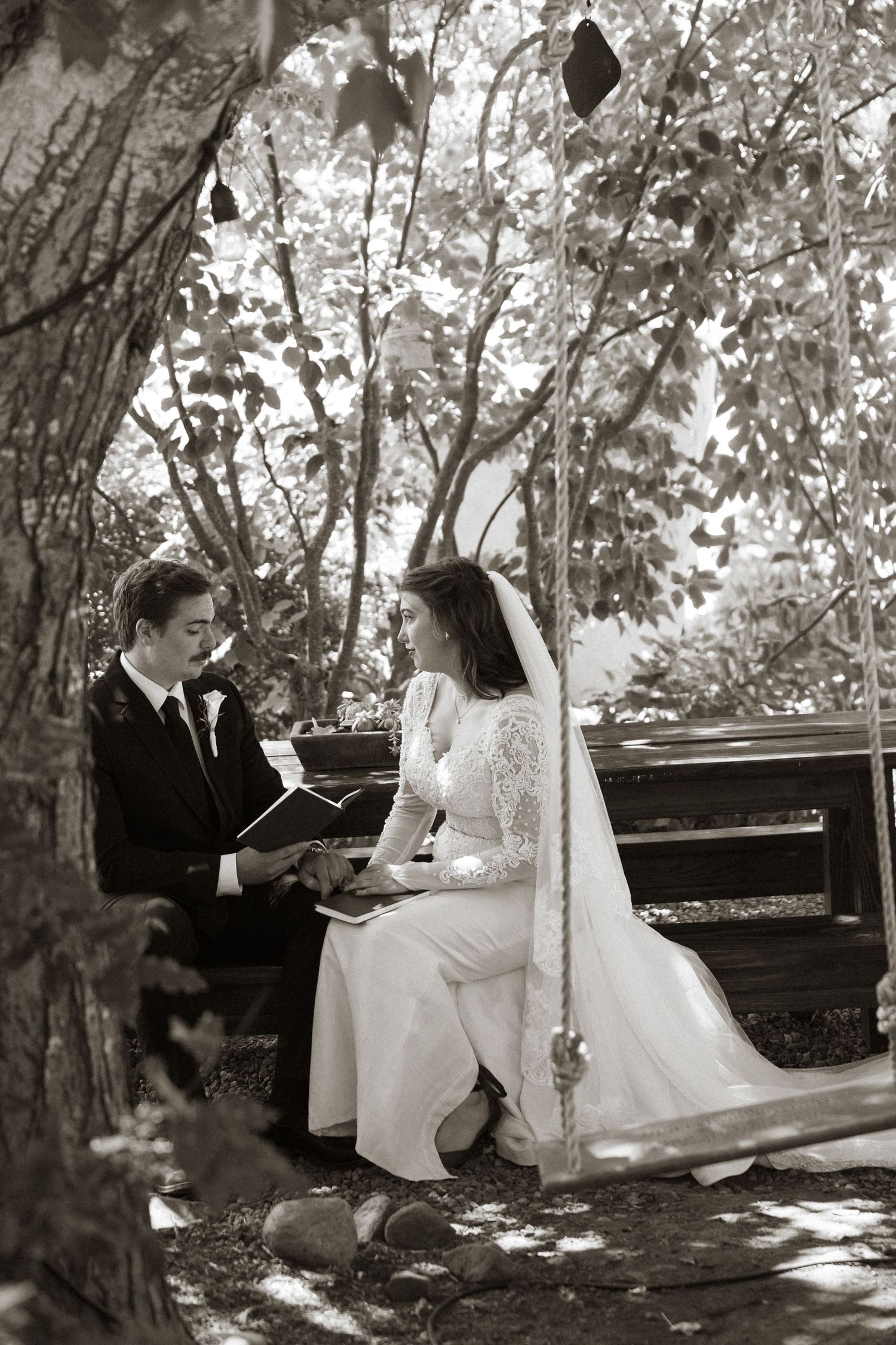 Black and white photo of bride and groom sitting on bench in garden, reading wedding vows together.