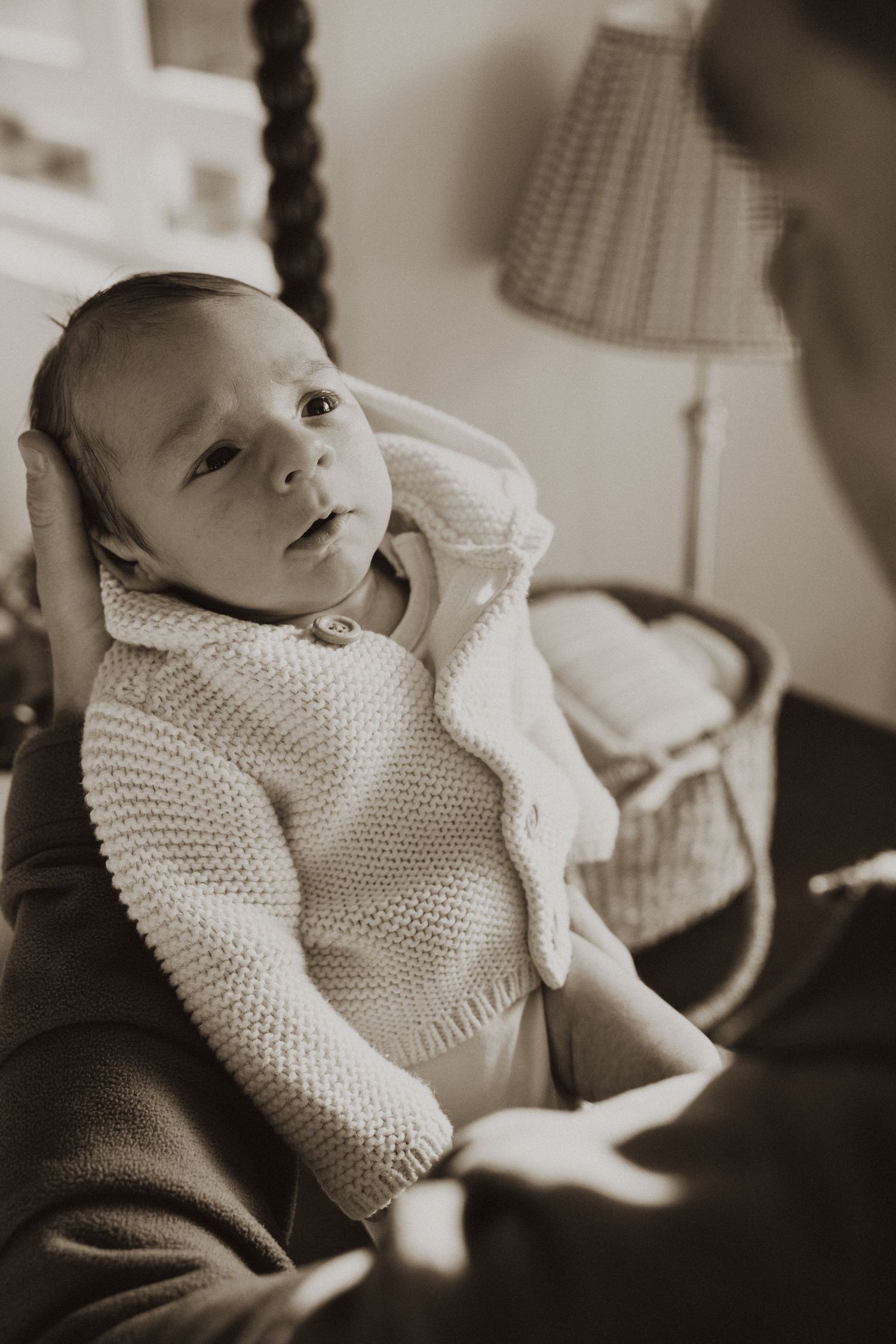 Newborn baby in knit cardigan being held by parent in sepia-toned nursery photo.
