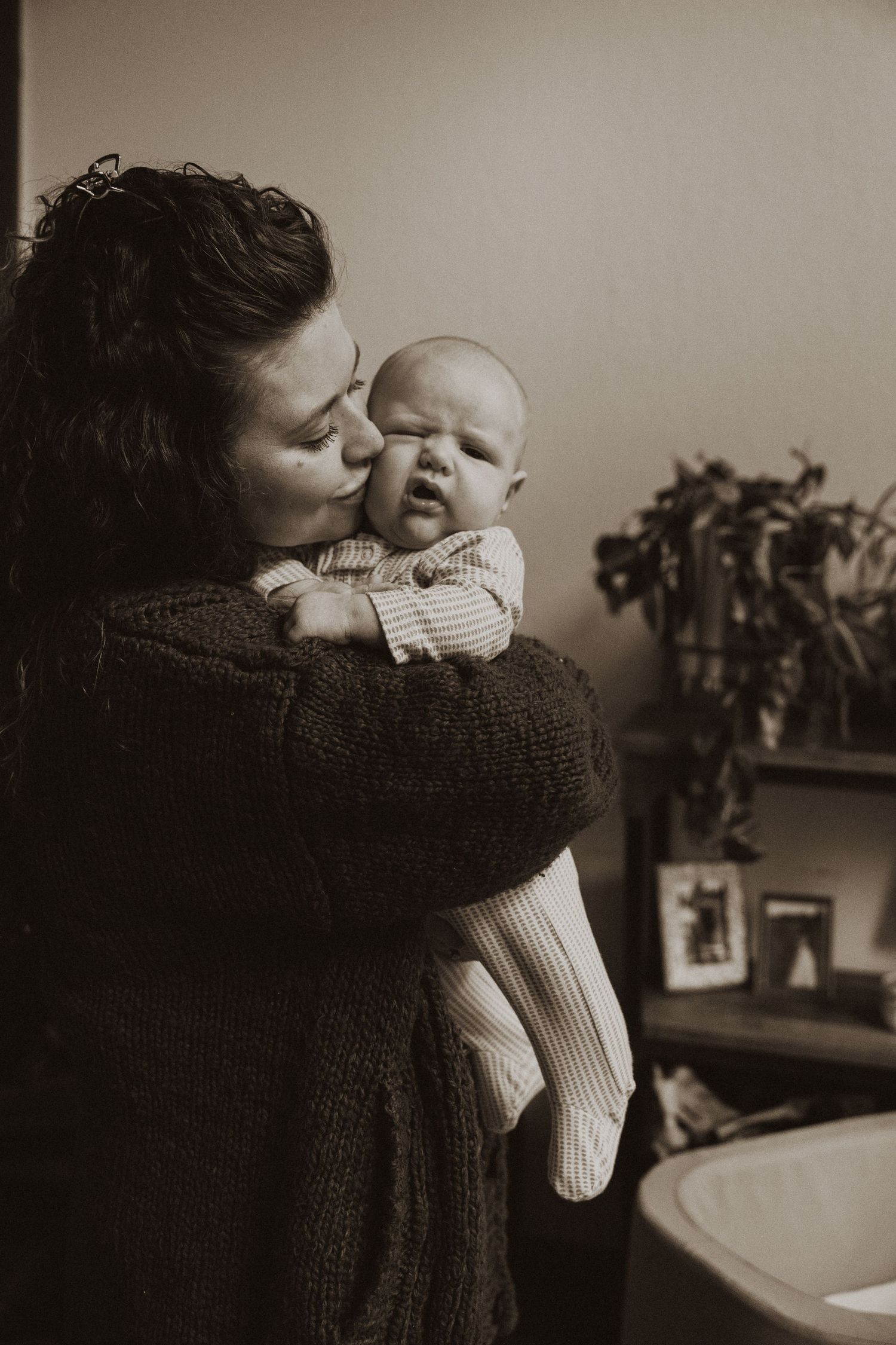 Mother kissing her newborn baby while holding them close in a warm sepia-toned portrait.